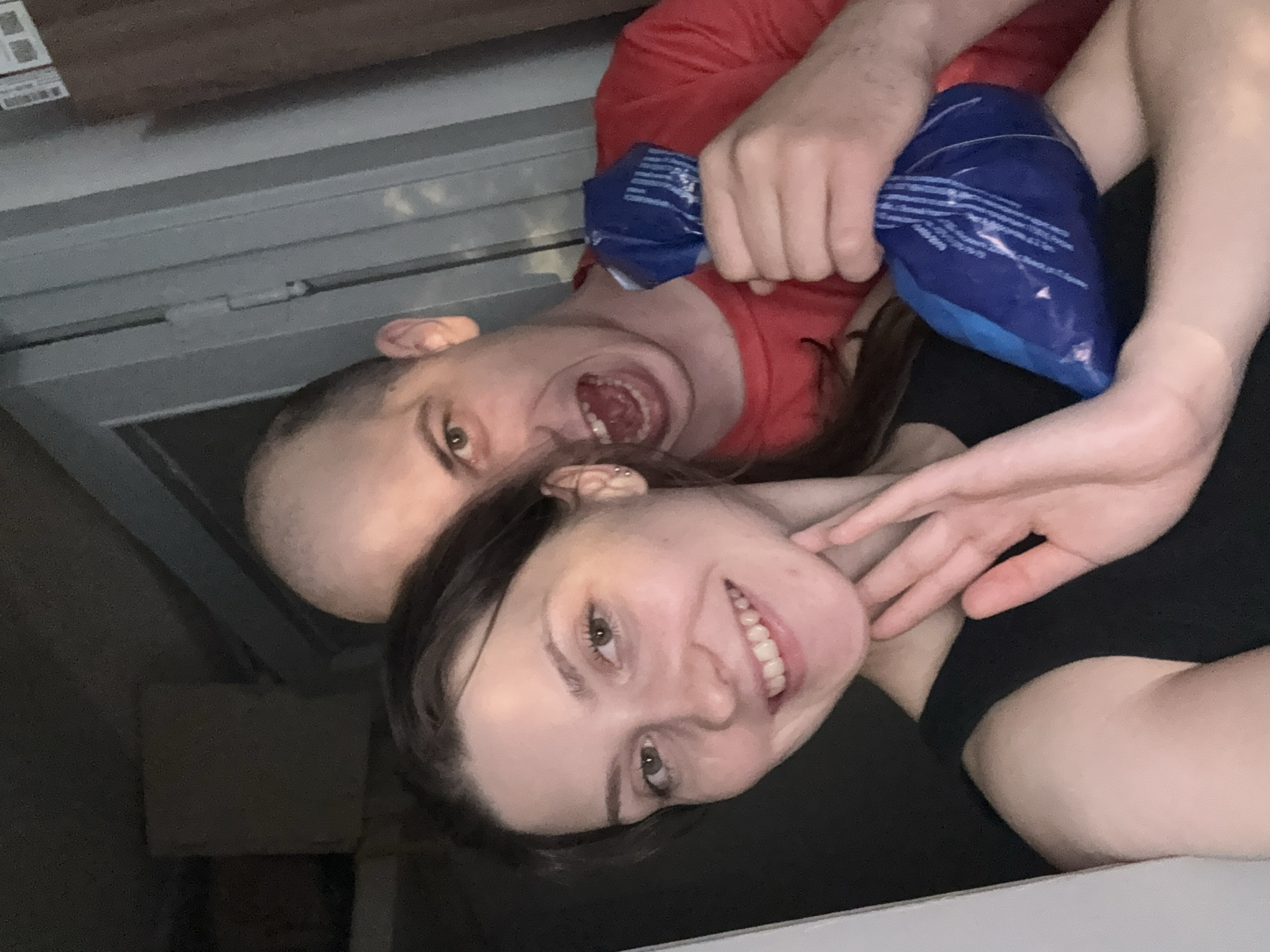 This image depicts a close-up, candid selfie of two individuals, a woman and a man, in what appears to be an indoor setting, possibly a residential hallway or doorway. The woman is in the foreground, smiling at the camera with her left hand raised, palm facing outward, near her chin. She is wearing a black sleeveless top. Her dark brown hair is pulled back. Behind her, the man is leaning forward, his face close to the camera. He has a bald head and is making a grimacing or shouting expression, with his mouth open wide. He is wearing a red t-shirt and holding a blue, flexible object, possibly an ice pack or a bag of frozen goods, in his right hand, which is positioned near the woman's shoulder. The background is dimly lit and appears to be a wall with doorframes or paneling. There are no specific identifiable landmarks or features that would indicate the location in Saint Petersburg, Russia.