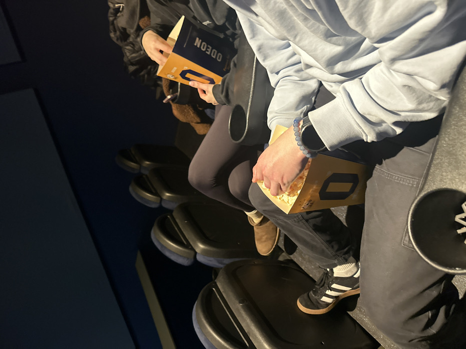 Inside a movie theater, two people sit side-by-side. One person wears a light blue hoodie and black sneakers with white stripes, holding an Odeon popcorn container. Their companion wears light brown boots and leggings, also holding a popcorn container. The Odeon logo is visible on both containers. The movie theater seats are black with blue accents. The scene is dimly lit, emphasizing the cinematic experience.