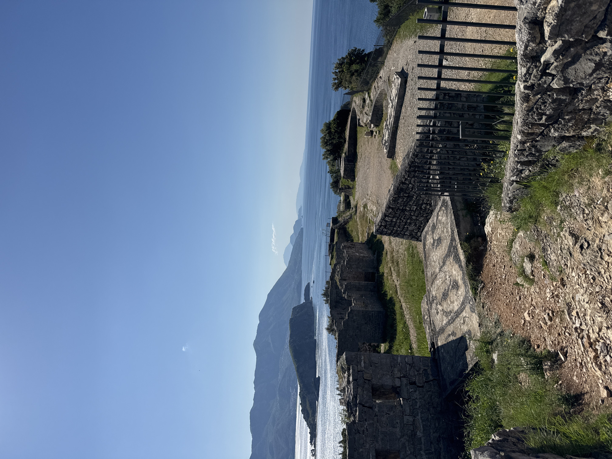 The image shows a coastal view from the ruins of a stone structure, possibly a fortress or castle, in the city of Budva, Montenegro. The view extends over the Adriatic Sea, with glimpses of distant mountains and islands. The ruins are characterized by weathered stone walls and open spaces, partially covered with vegetation like grass and small trees. Modern additions include metal fencing along the perimeter of the ruins. The sky is clear and blue.