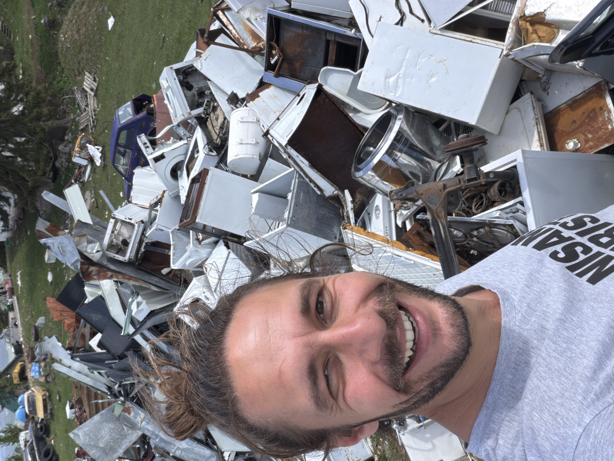 A smiling man is taking a selfie in front of a junkyard. The junkyard is full of metal debris, including discarded appliances and vehicle parts. The junkyard appears to be outdoors, with a grassy area and scattered buildings visible in the background. It is not possible to determine if this is in Žabljak, Montenegro, without additional information.