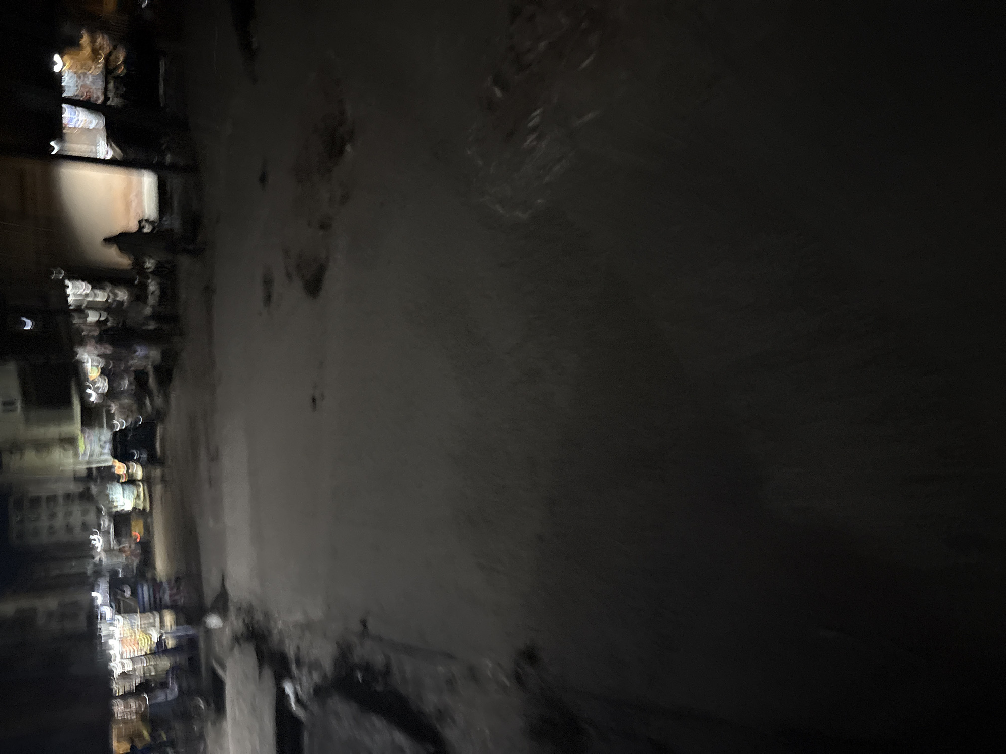 The image depicts a street scene at night in Onitsha, Nigeria. The overall scene is blurry due to motion, suggesting movement of the camera or subjects. In the background, there are buildings illuminated by artificial lights, indicating an urban environment. There appear to be several vehicles, possibly including cars and motorcycles, with their lights visible as streaks of light. Multiple individuals are present in the scene, some walking and others standing near what appear to be stalls or shops. The street itself is paved and appears wet in some areas, reflecting the lights. The atmosphere is dark, characteristic of nighttime.