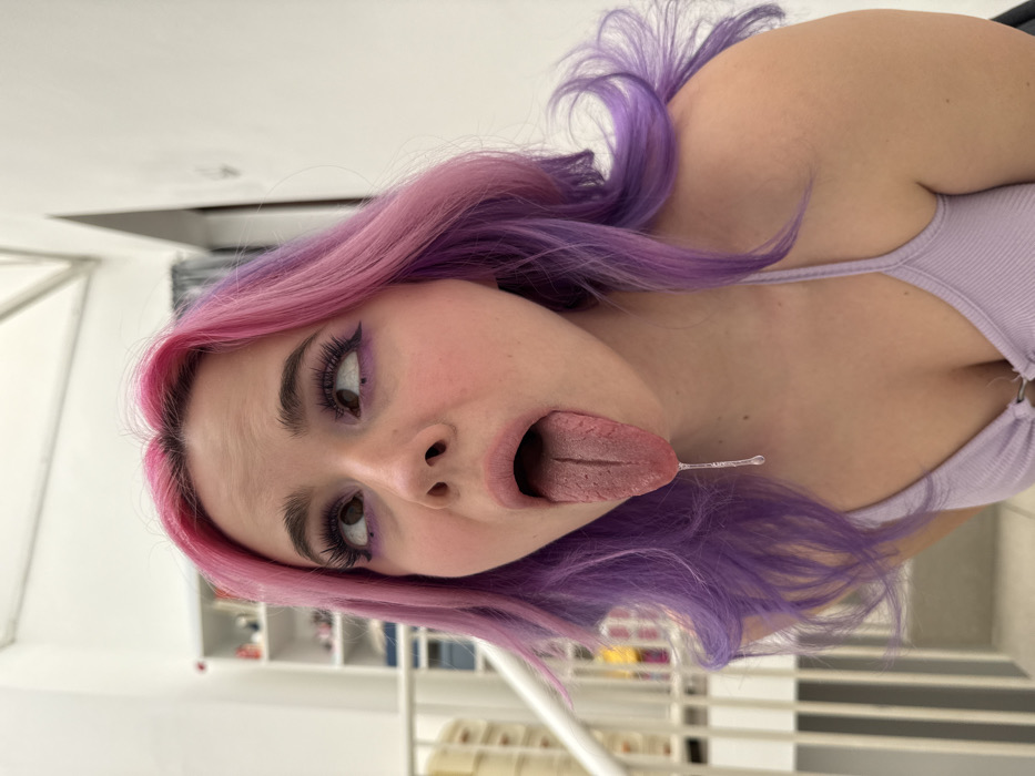 A young female is depicted in a close-up, indoor portrait shot in the City of Zagreb, Croatia. She has light skin, long wavy hair dyed bright pink at the roots transitioning to purple, and wears a light purple ribbed tank top. Her eyes are rolled upwards and slightly to the right, with prominent dark purple winged eyeliner and false eyelashes. Her mouth is open wide, with her tongue fully extended downwards, from which a single strand of saliva drips vertically. In the background, to the left, a white staircase railing is visible, along with a partial view of a white shelving unit containing various items. The walls in the background are plain white, indicative of a modern interior space.