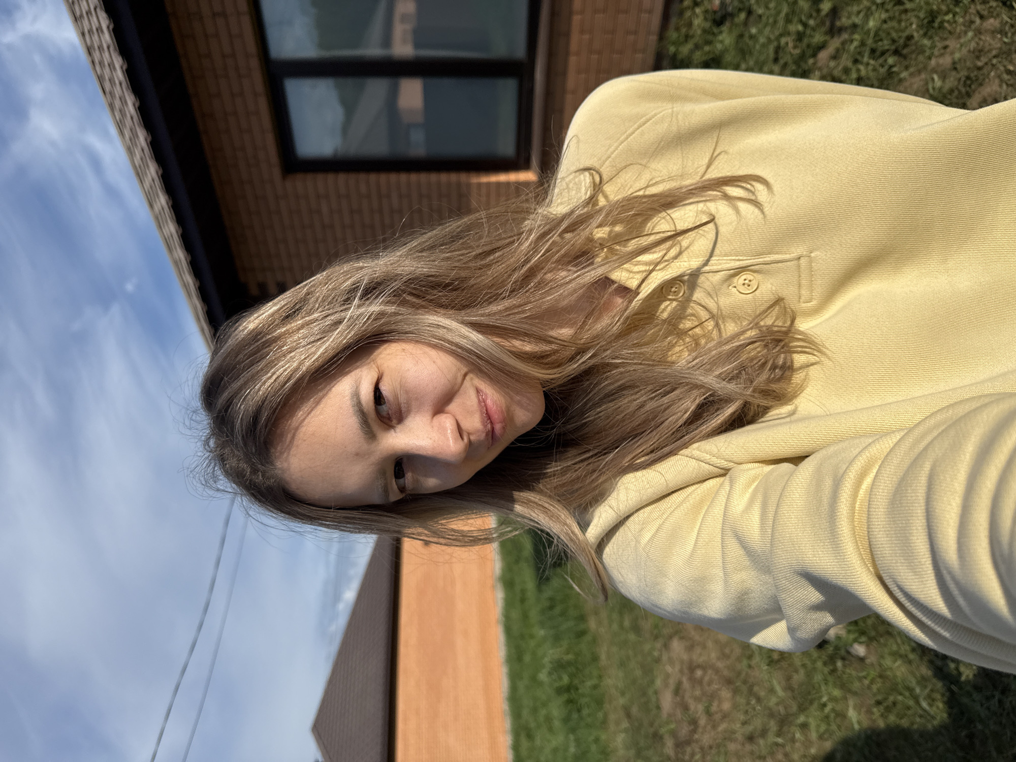 A young woman with blonde hair is positioned in the center of the image, wearing a yellow collared shirt. She is tilting her head and smiling slightly.
Behind her is a brick building with a window. To the left, there is a blue sky and the corner of another brick building. Green grass is visible along the right side and bottom of the frame. A powerline is seen against the sky in the lower left.
The location is outdoors in Chaikovsky, Russia.