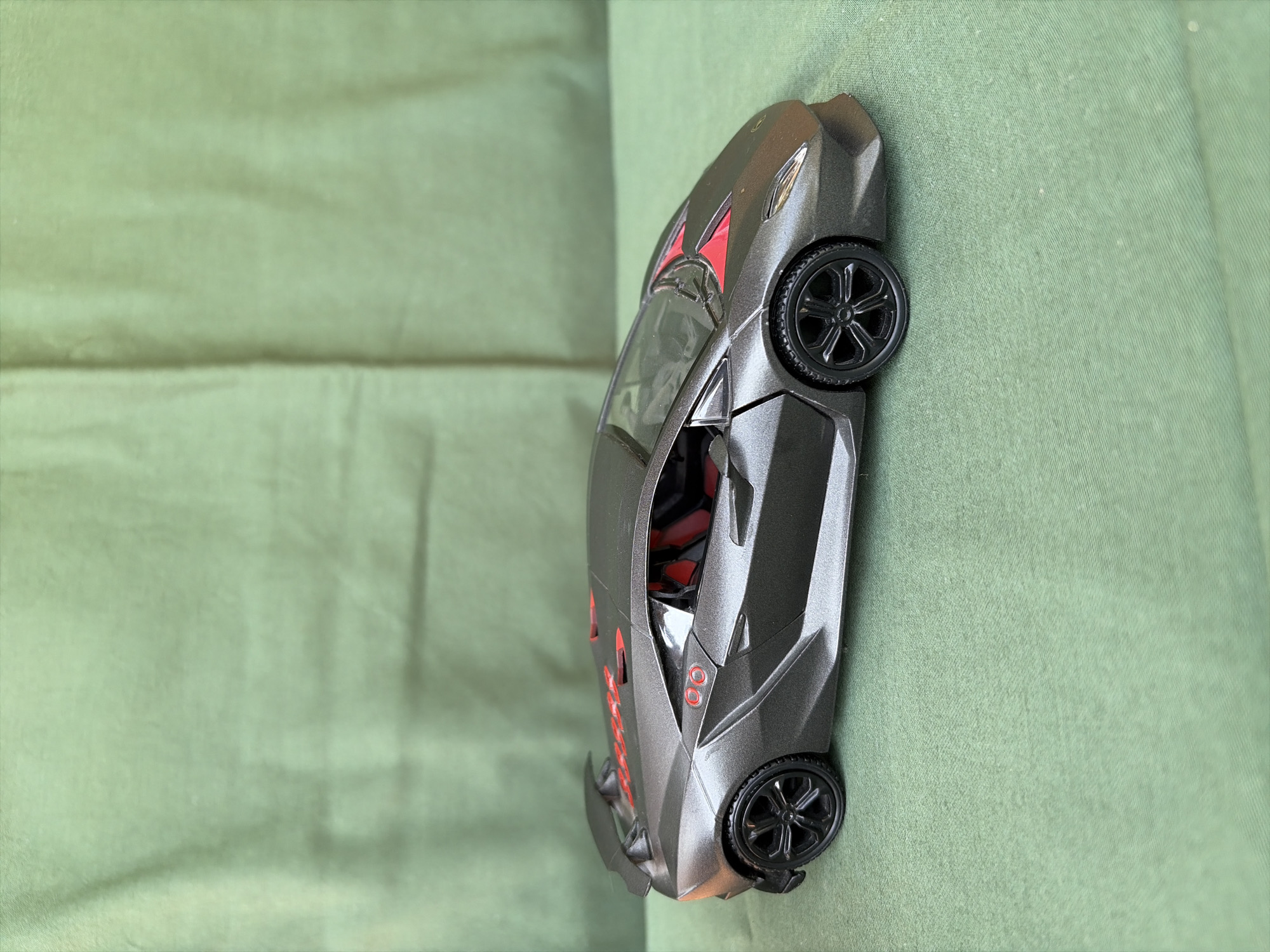A gray Lamborghini toy car with red accents sits on a green, textured surface. The car is viewed from the side, showing its sleek design, including a spoiler and black wheels. The background is a solid green, slightly wrinkled fabric. There are no people or any other objects visible in the image. The scene is static and appears to be a product photograph. The city of Oxentea, Moldova, is not depicted in this image.