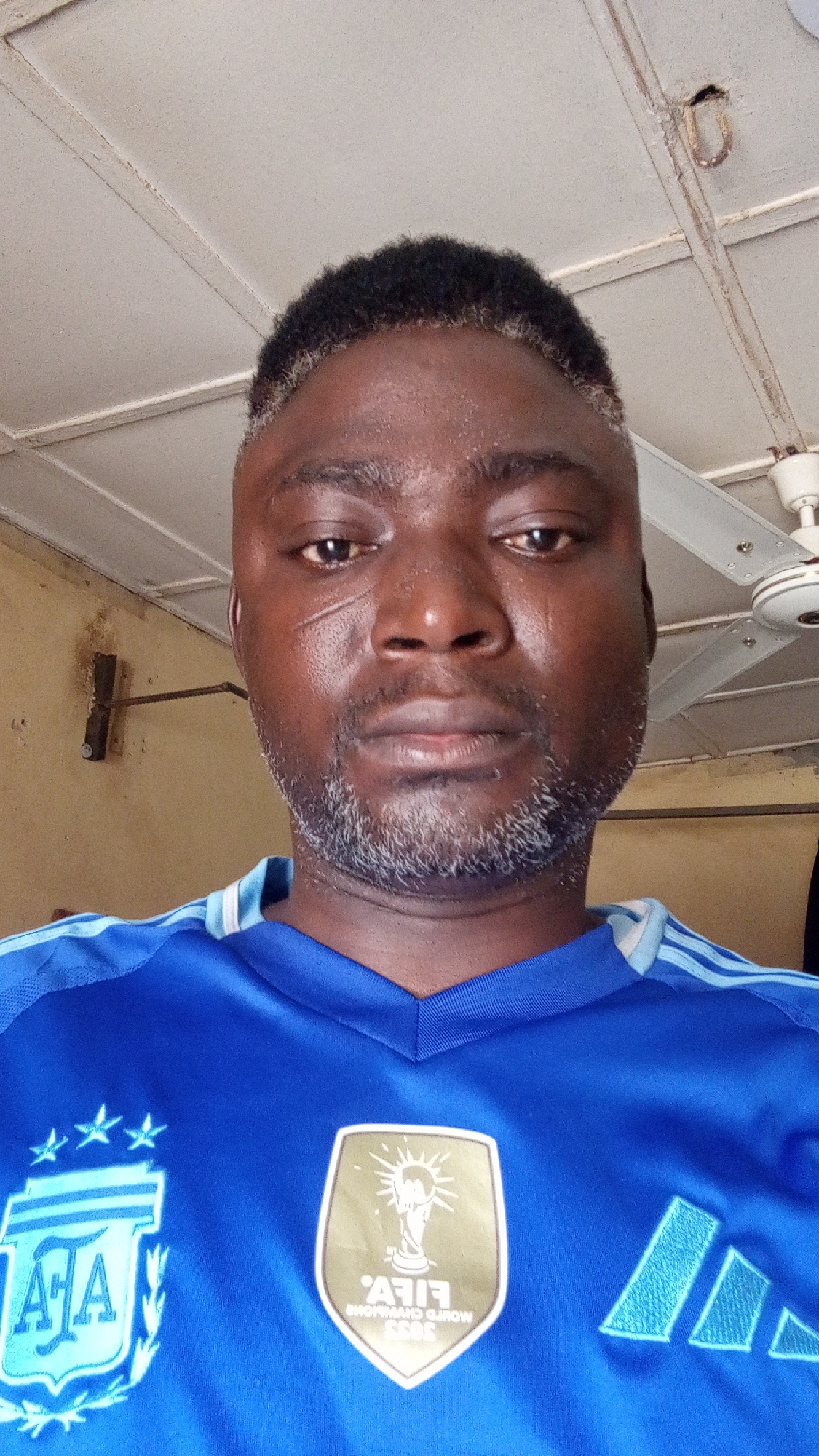 A dark-skinned man with dark hair and a graying beard is shown in a close-up, selfie-style shot. He has a scar on his left cheek. He is wearing a blue Argentina soccer jersey with the team's crest and the FIFA World Cup patch. The background shows a ceiling with visible pipes and a ceiling fan. The location is Sabon Tasha, Nigeria.