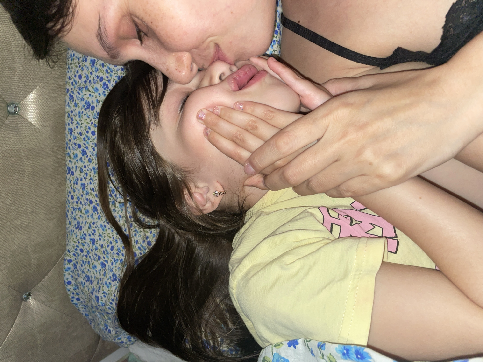 A woman is kissing a child. The woman has dark hair and fair skin. She is wearing a black bra. The child has dark hair and fair skin. They are lying on a bed with a floral patterned sheet. The child is wearing a yellow shirt with a pink design. A hand is covering the child's face. The location in the city of Izhevsk, Russia is not discernible from the image.