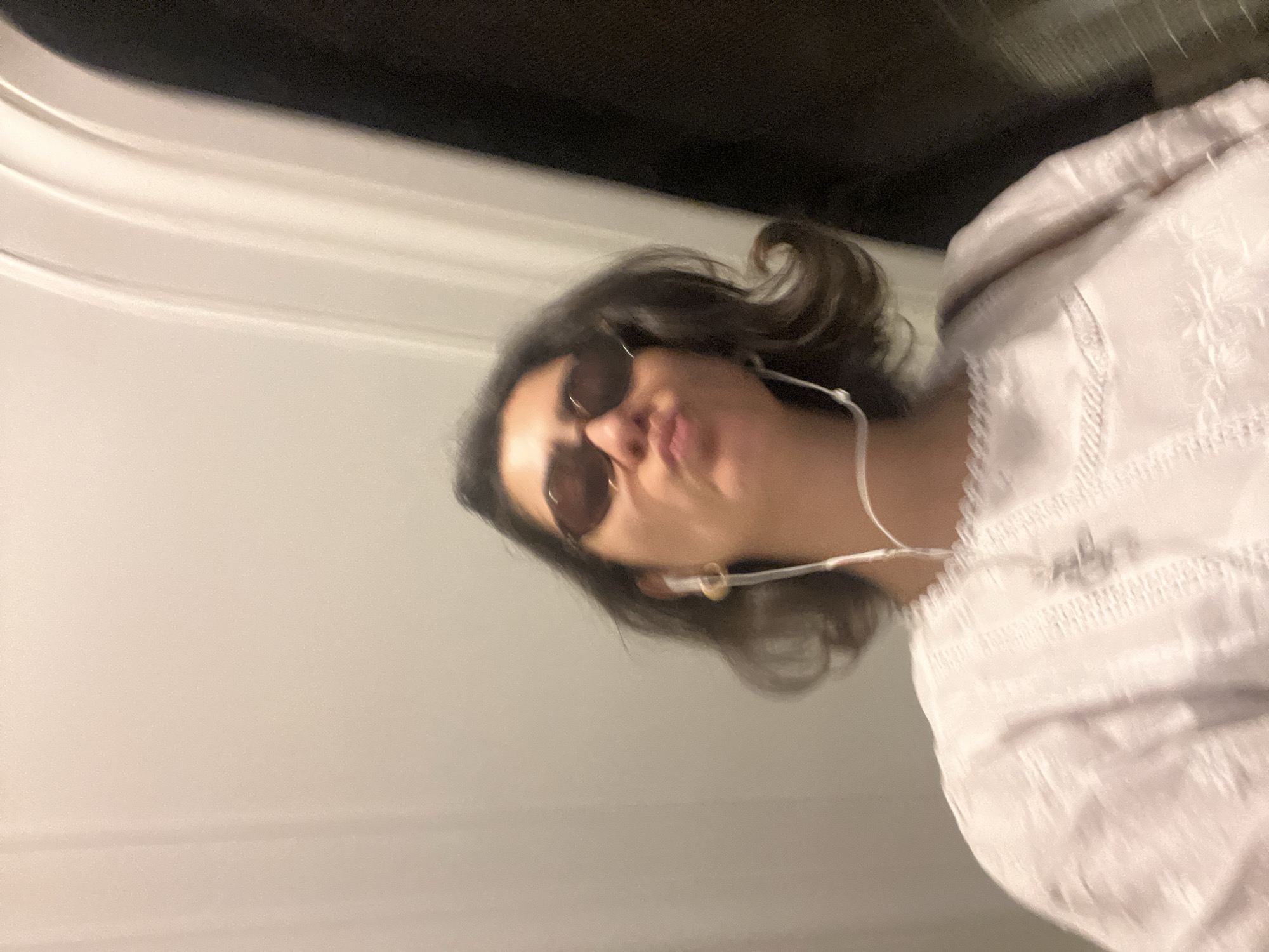 A woman with dark hair and sunglasses is positioned in what appears to be an indoor setting, possibly a train or subway car based on the white walls and ceiling visible in the background. She is wearing a white shirt with embroidered details and has earphones in. The photograph is taken from a low angle, slightly tilted, giving a perspective that looks up at the woman. Based on the background, this could be a subway train in the city of Paris, France.