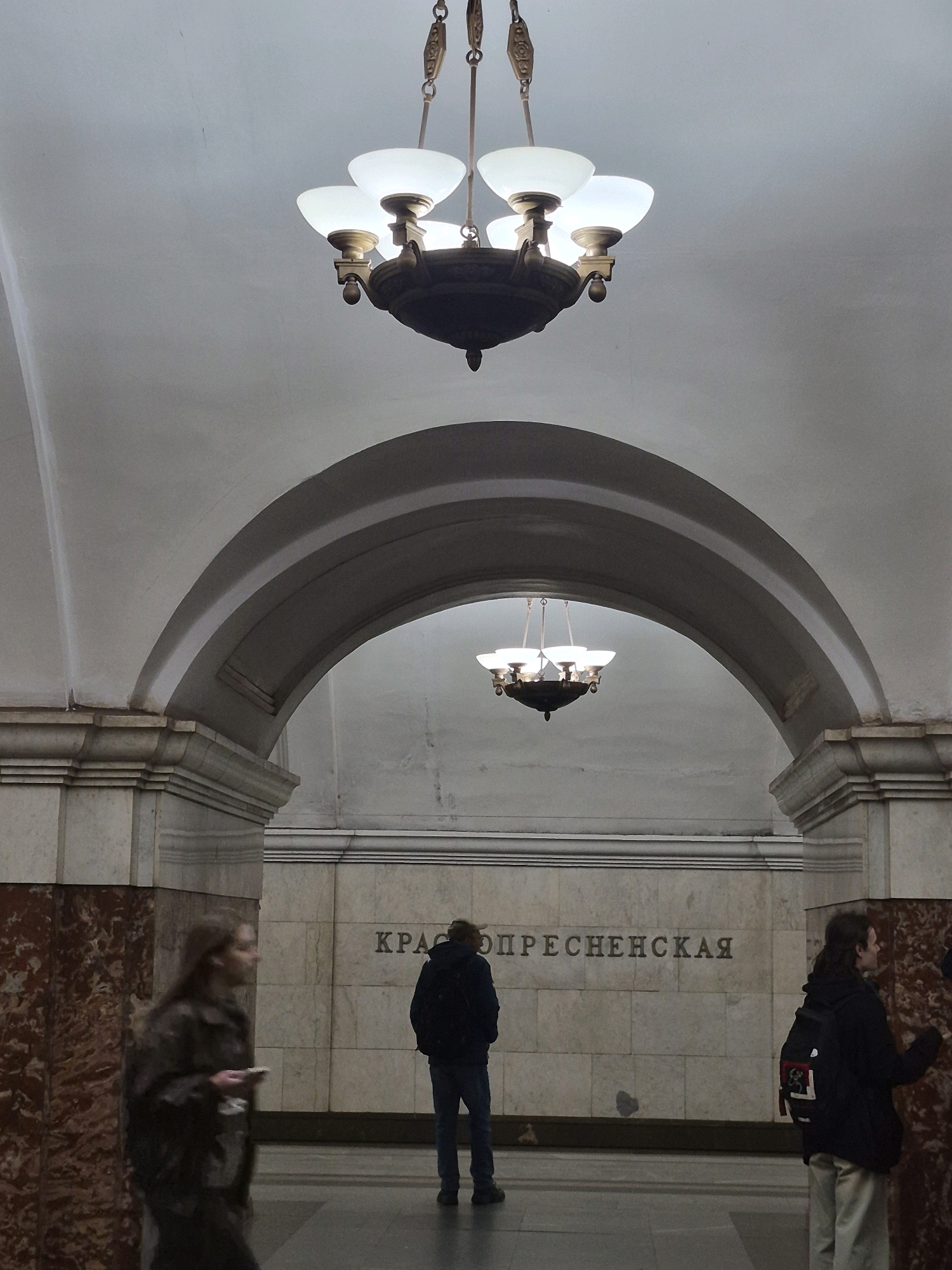 The image shows an interior scene of a subway station with arched ceilings and ornate chandeliers. In the foreground, a person with long brown hair walks to the left, holding a smartphone. To the right of the person, a man wearing a dark jacket and backpack stands facing the wall with Cyrillic lettering that reads "КРАСНОПРЕСНЕНСКАЯ". Further to the right, a person in a black jacket and light-colored pants stands facing the wall. The walls are made of marble and textured stone. The lighting is dim and casts shadows, highlighting the architectural details of the station.