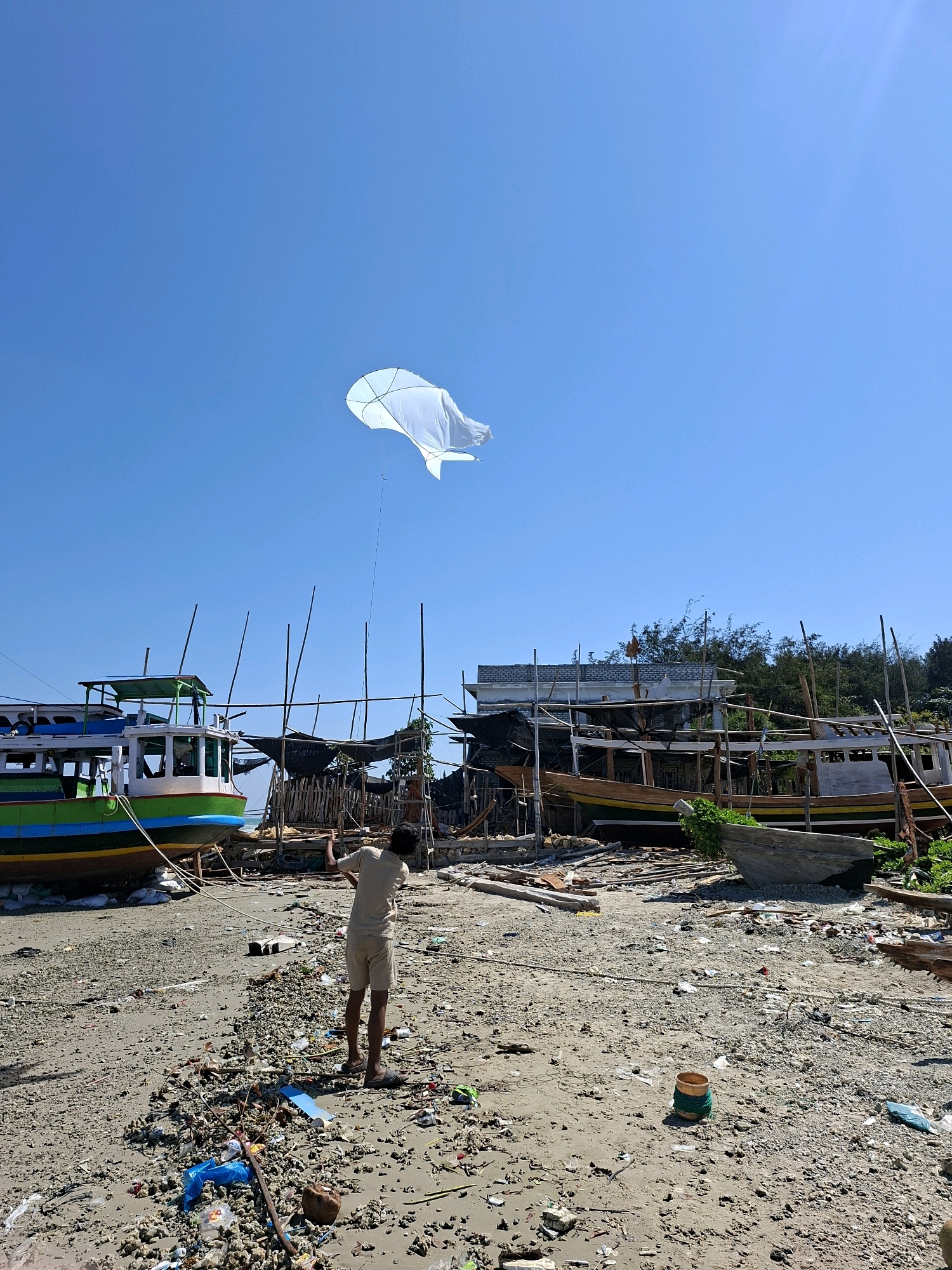 The photograph depicts a beachside scene in Camplong, Indonesia, characterized by a clear blue sky and a sunny day. In the foreground, a person wearing a light-colored t-shirt and shorts stands facing away from the camera, holding the string of a large, white kite that is airborne in the upper portion of the frame. The beach itself is covered with sand, debris, and various objects, including pieces of wood, plastic waste, and a small wooden bucket.
The midground of the image features several boats, some of which appear to be under construction or repair, situated on the sand. One boat in the left foreground is brightly colored with green, blue, and yellow stripes. Behind these boats, there is a makeshift structure made of wood and netting, suggesting a boatyard or maintenance area. Trees and vegetation are visible in the background, separating the beach from the sky. The overall environment appears to be a working area for boat maintenance and construction, with a strong presence of both maritime activity and litter.