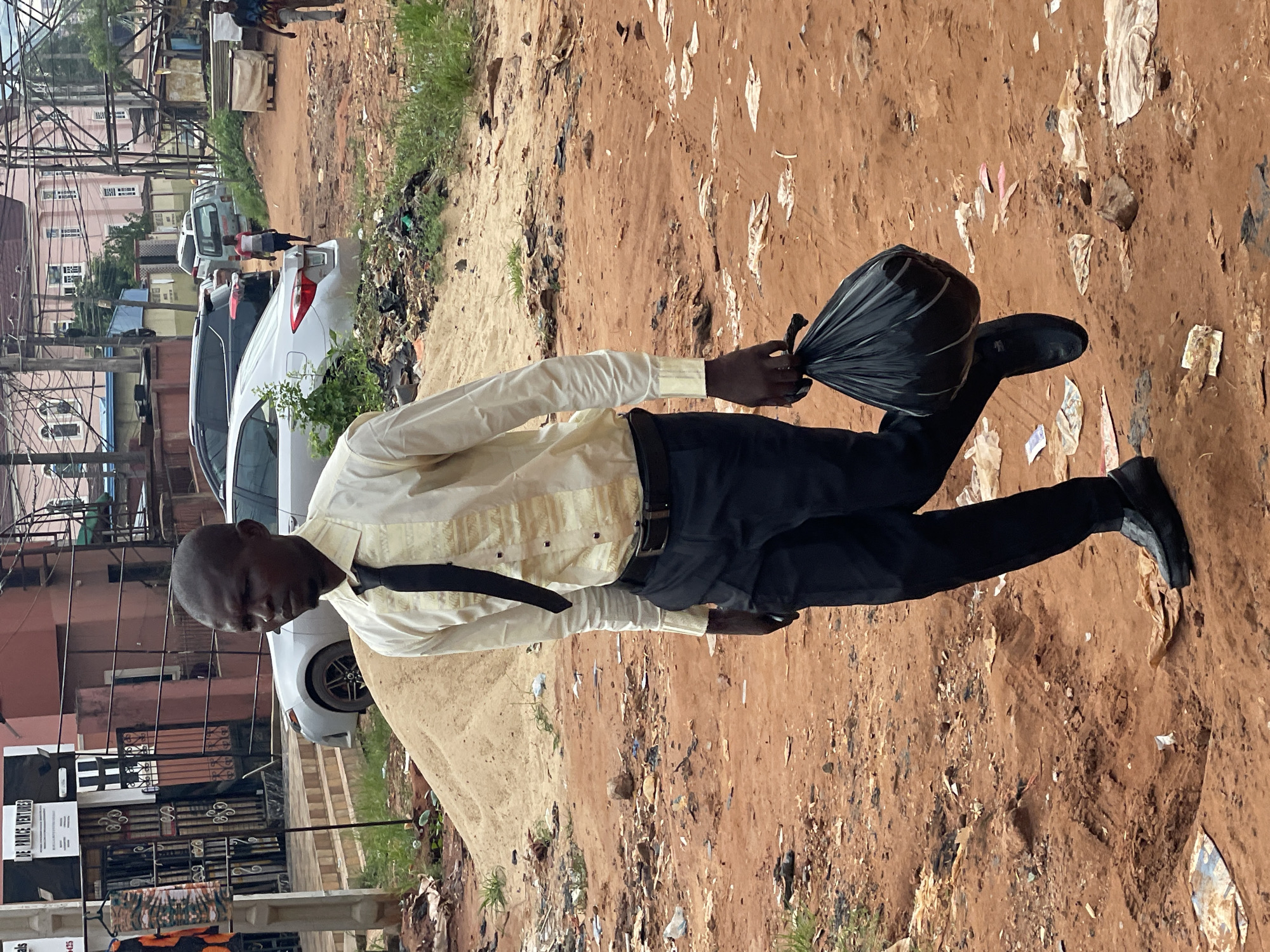 A man dressed in a long-sleeved yellow shirt, black pants, black shoes, and a black tie walks along a dirt path in Nkpor, Nigeria. He is carrying a black plastic garbage bag in his left hand. The path is strewn with litter. A white car is parked along the side of the path. Buildings, power lines, and other cars can be seen in the background.