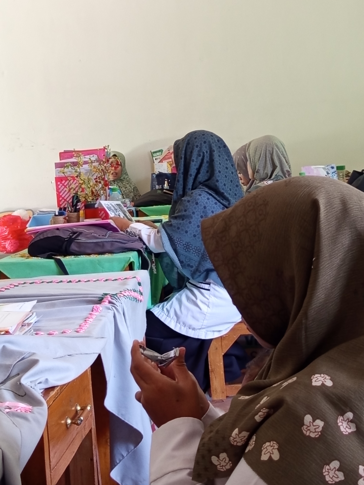 In Lumutan, Indonesia, a group of women, identifiable by their hijab head coverings, are seated at tables in a classroom or meeting room. The tables are covered with light-colored cloths decorated with pink ribbon trim. On the tables, there are books, folders, pens in a holder, a vase with artificial flowers, and a bottle of water. One woman in the foreground is in profile, wearing a brown hijab with a floral pattern, and appears to be holding a nail clipper. Behind her, another woman in a dark blue hijab is visible. Further back, a woman in a green headscarf is seated at a table, and other individuals are partially visible. The room has plain, light-colored walls. The overall impression is one of a study session or a group activity within an educational or community setting.