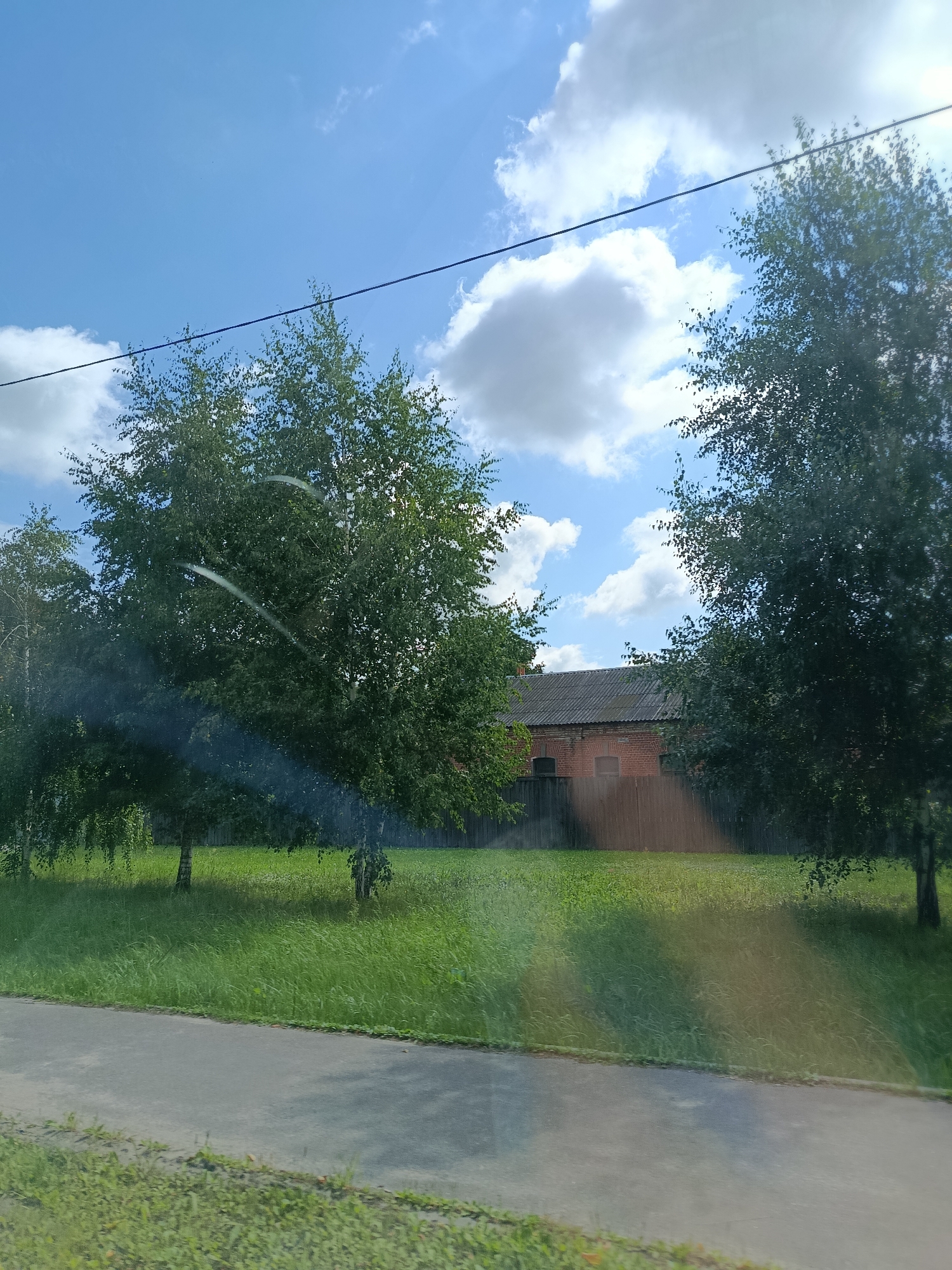 The image depicts a sunny day in Orekhovo-Zuyevo, Russia, with a clear blue sky dotted with fluffy white clouds. A power line runs horizontally across the upper portion of the frame. In the foreground, a paved path runs alongside a lush green grassy area. Beyond the grass, a low wooden fence separates the yard from a long, low-rise building constructed of red brick. The building has a dark, corrugated roof and several small, dark-framed windows. Two prominent birch trees with green foliage are situated in the middle ground, casting shadows on the grass. The overall scene suggests a quiet, rural or suburban setting.