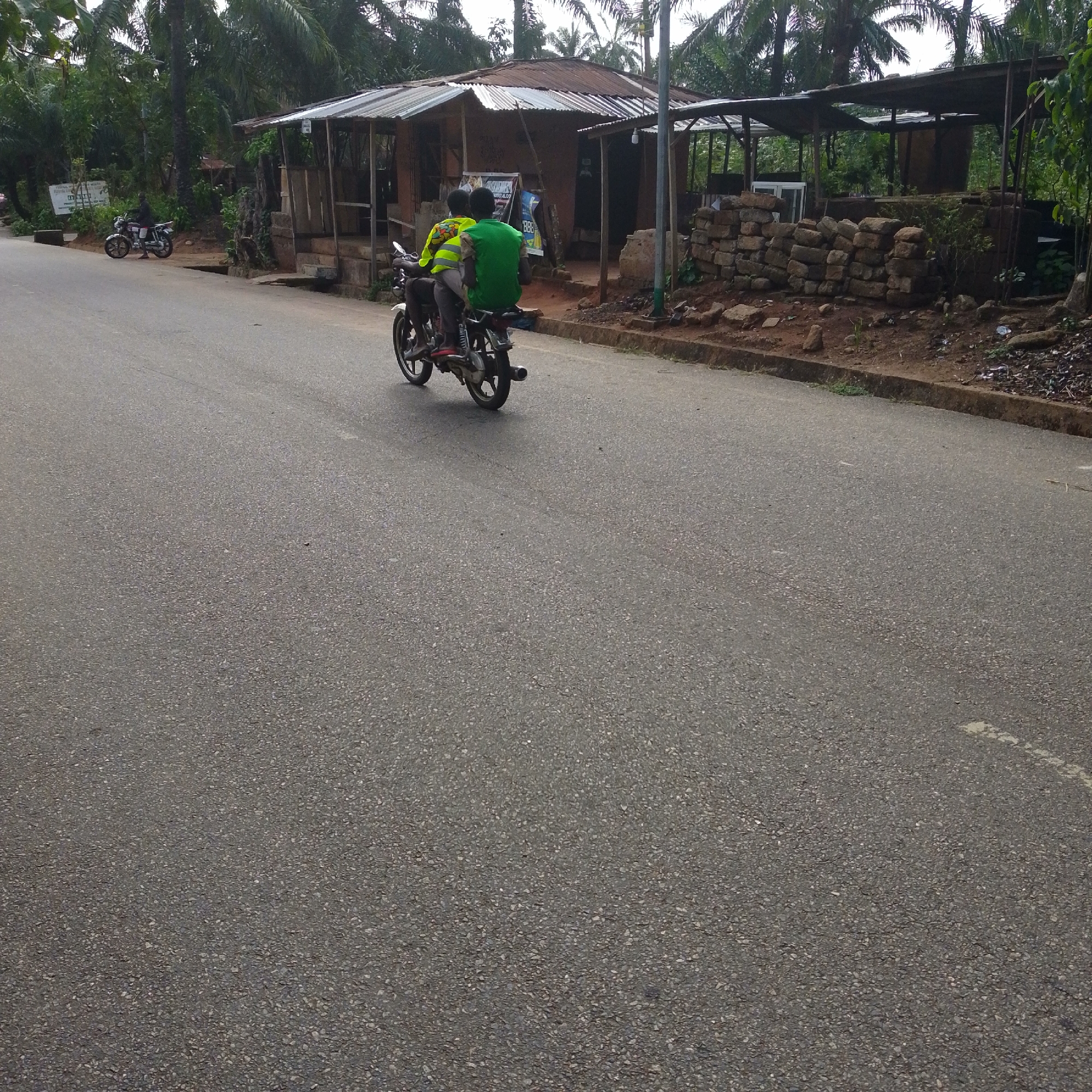 Two individuals are riding a motorcycle on an asphalt road in a tropical, rural environment. The person in the front is wearing a green shirt, while the person behind is wearing a bright yellow jacket with reflective strips. The motorcycle is dark-colored with white accents.
The location includes simple roadside structures, some made of stone and wood, with corrugated metal roofs. Palm trees and other tropical vegetation are visible in the background. The road appears to be in a location with limited infrastructure. Another motorcycle is parked on the side of the road in the distance.