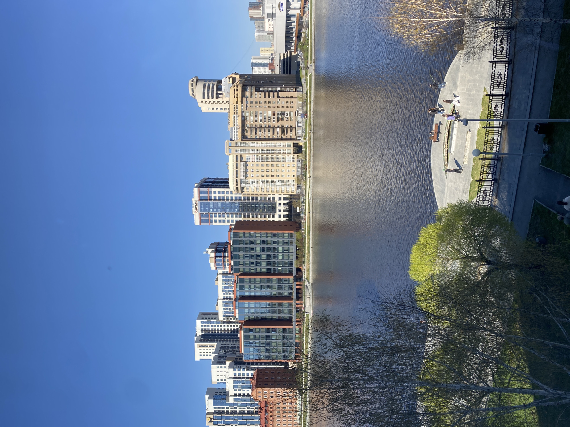 The image depicts a waterside scene in Yekaterinburg, Russia, featuring modern buildings along a riverbank. Clear blue sky is present. The buildings range from red brick to white and glass structures. A body of water reflects the buildings and sky. Alongside the river, there's a paved area, possibly a walkway or park, with trees in early spring foliage. People are standing and sitting near the river. Black decorative fence is visible.