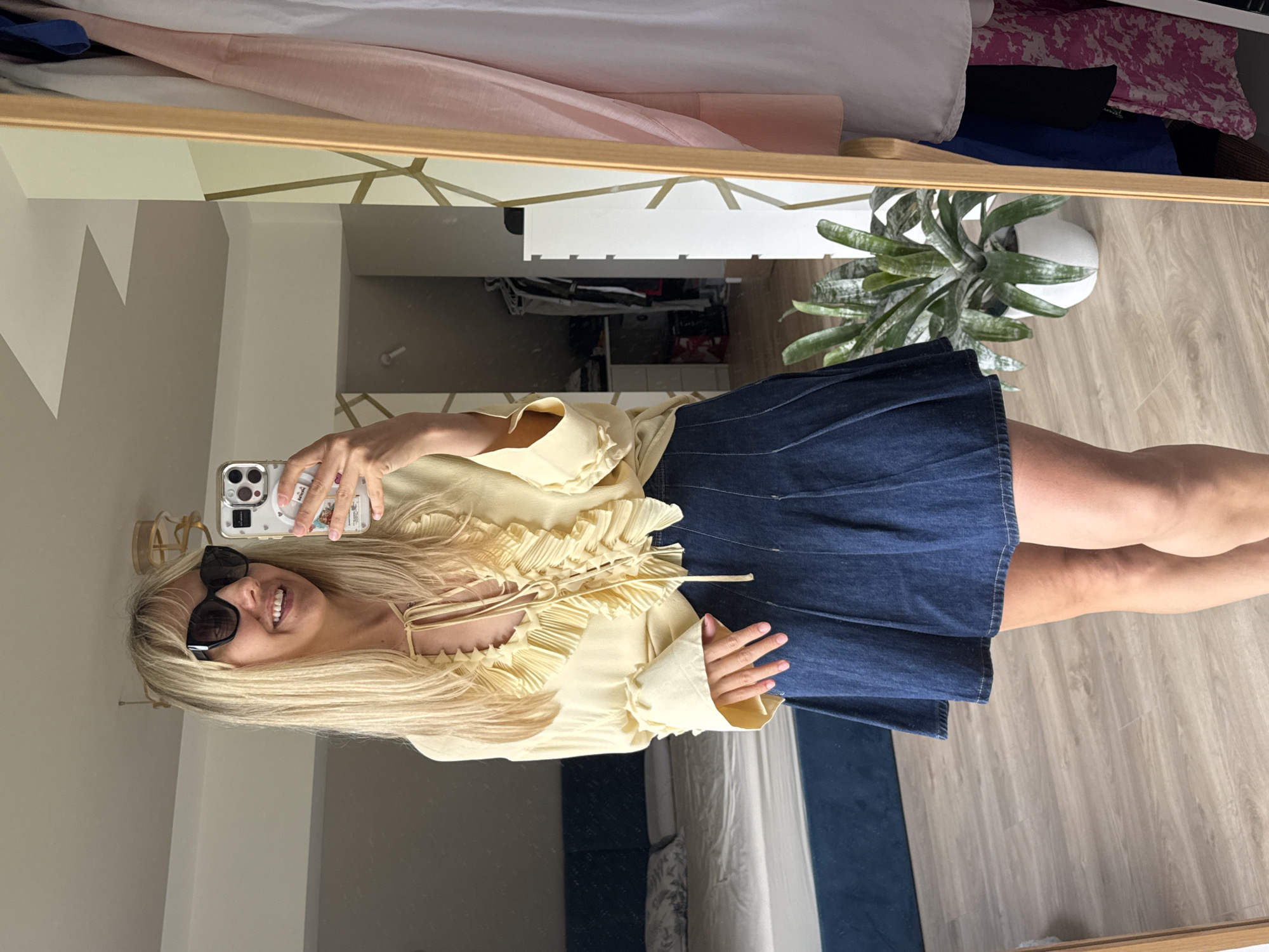 A woman is taking a mirror selfie in a bedroom. She has blonde hair and is wearing a light yellow ruffled blouse and a dark denim skirt. She is also wearing sunglasses and smiling. In the background, there is a bed with a pink and white patterned duvet, a closet with clothes, and a potted plant. The floor is made of light wood laminate. The room appears to be well-lit, suggesting it is daytime. The overall mood of the image is casual and relaxed. There is no visible text in the image.