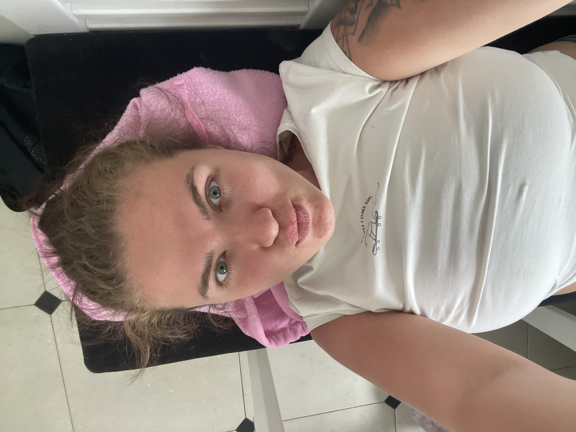 A person is lying on a black surface, possibly a table or mat, with a pink towel or cloth partially under their head. The person is wearing a white t-shirt. A tattoo is visible on the upper arm. The background includes white tile with some black accents. The location cannot be determined based on visual information.