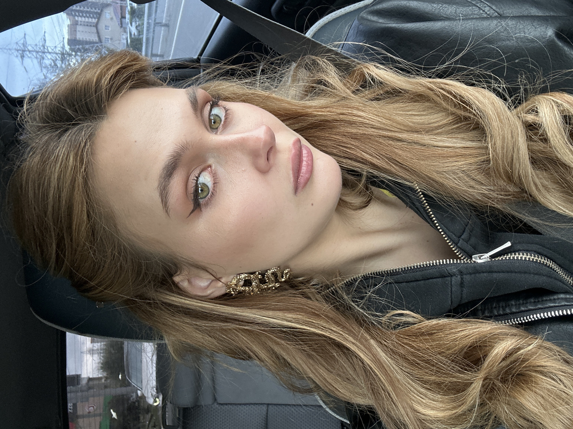 The image is a close-up, selfie-style portrait of a young woman with long, light brown hair and green eyes. She is wearing a black leather jacket and a gold-colored earring shaped like the word "Boss". The photo appears to have been taken inside a car, as evidenced by the seatbelt across her chest and the car window reflecting a cityscape. The background suggests a location in the city of Krasnoyarsk, Russia. A view of the buildings in the distance supports this.