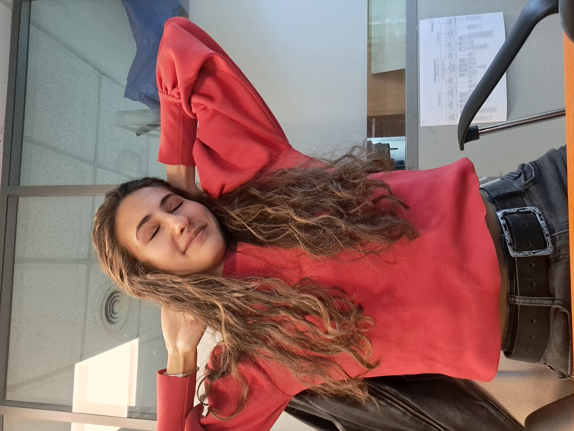 A young woman with long, wavy, light brown hair is depicted indoors, likely in an office or modern building, under natural daylight. She is seen from the chest up, resting with her eyes closed and a soft, contented smile on her face. Her posture suggests she is leaning back in a chair, with her right arm bent and her hand gently resting near her head. She is wearing a loose-fitting, long-sleeved red top and dark grey jeans with a black belt featuring a decorative silver buckle. A silver bracelet adorns her right wrist.
The background includes a large glass partition or window to her left, reflecting a blue object and the textured ceiling. A strong rectangular highlight from sunlight is visible on this glass surface, indicating a bright daytime setting. To her right, a light-colored wall displays a document, possibly an organizational chart or detailed diagram, with various interconnected boxes and faint text, including "KPP-101" at the top. Part of a black chair is visible in the foreground to her right. The ceiling features standard acoustic tiles and a circular air vent. The overall impression is one of peaceful relaxation within a contemporary, professional environment in Sofia, Bulgaria.