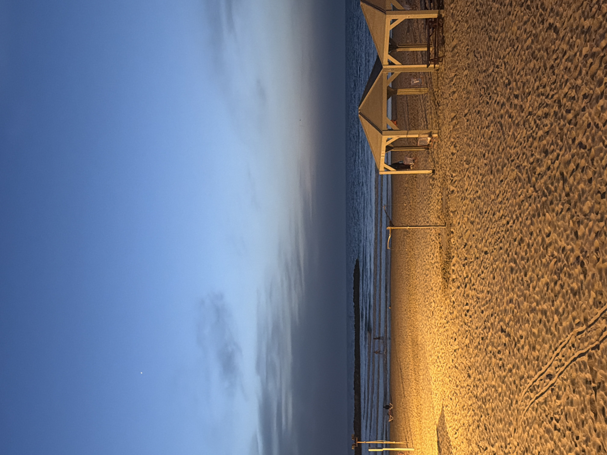 The image depicts a beachfront scene in Tel Aviv, Israel, at dusk. The sky is a gradient of blue to gray, meeting the sea. A line of identical, simple white structures resembling canopies or shelters is positioned along the beach. The sand is a light brown color and appears coarse, with visible footprints and tracks. The water is calm, reflecting the subdued light of the sky. The scene is lit mainly by artificial lighting from an unseen source, casting strong highlights on the sand. There are indications of people or objects under some of the canopies. The overall atmosphere is tranquil.