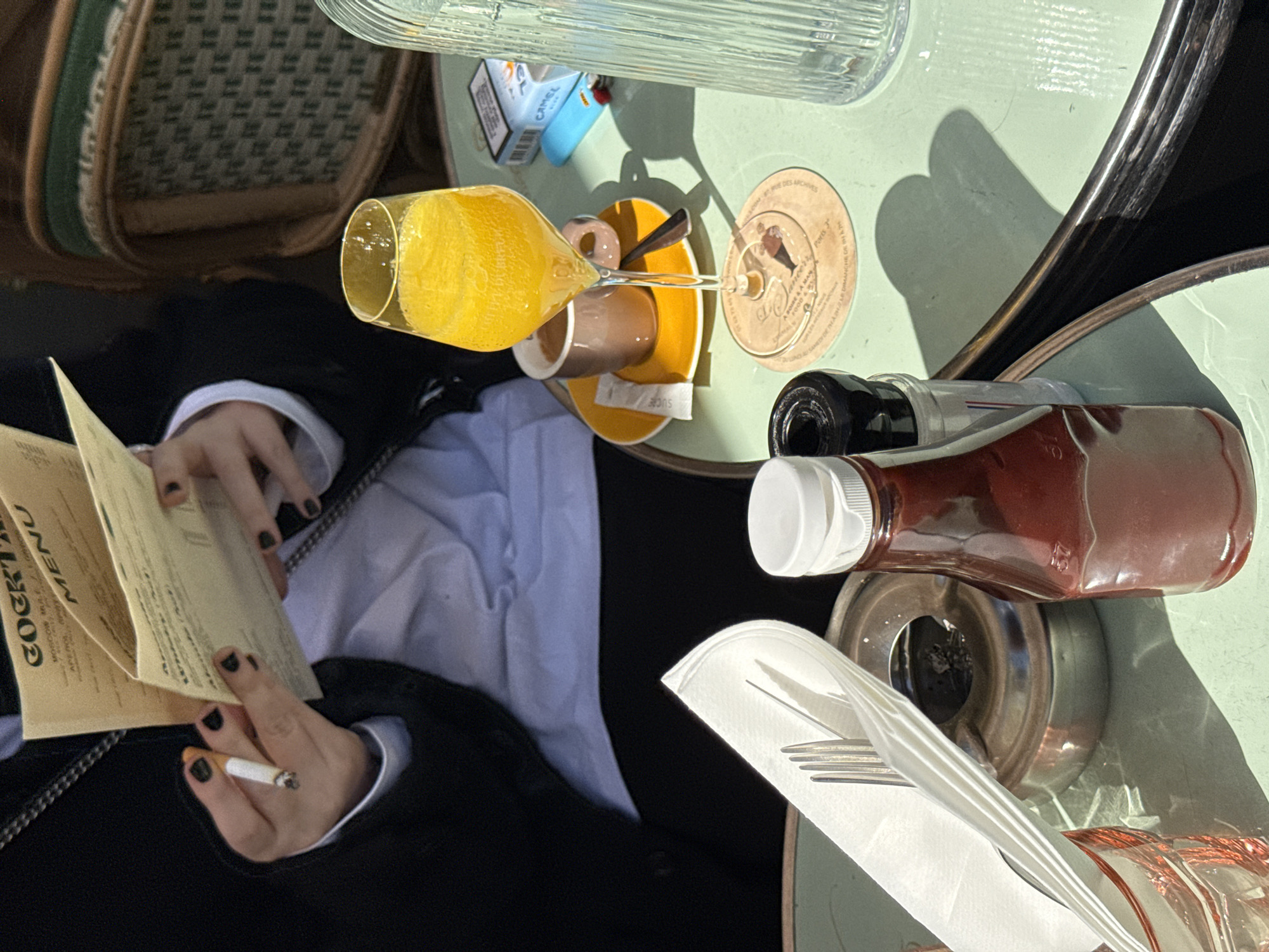 The image depicts a scene at an outdoor café, likely in Paris, France. A person is seated at a light green table, looking at a menu labeled "Cocktail Menu." The person is holding a lit cigarette in their left hand and has black nail polish. A glass of orange juice is placed on a yellow saucer next to a cup of coffee. A sugar packet labeled "SUCRE" is near the saucer. Other items on the table include a clear glass bottle of water, a pack of "Camel" cigarettes, a lighter, a coaster, a bottle of what appears to be ketchup, cutlery wrapped in a napkin, and an ashtray. A green and brown wicker chair is partially visible to the left.
