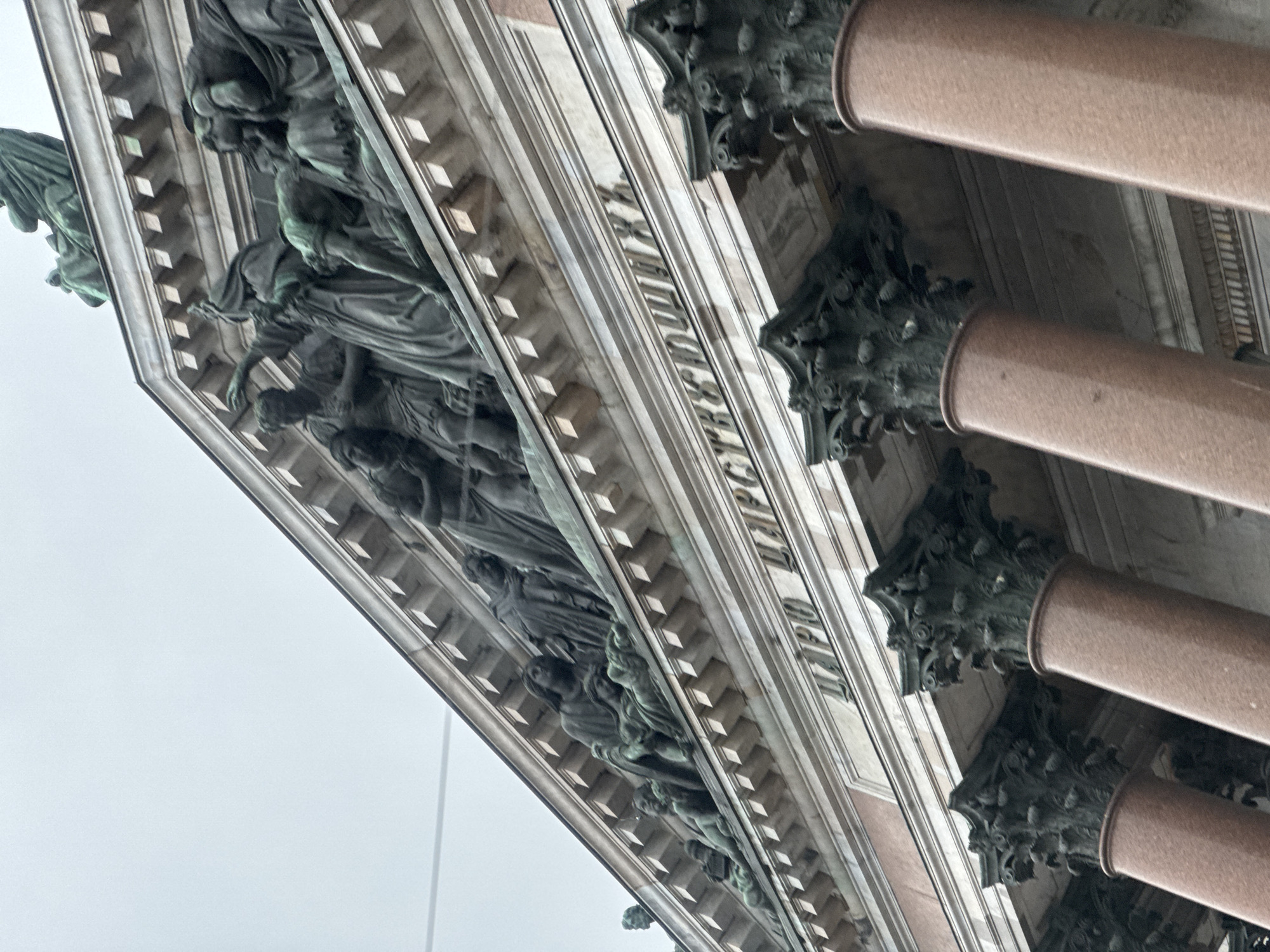 The image displays a close-up, low-angle view of a monumental architectural facade, rotated 90 degrees counter-clockwise. The scene is dominated by classical architectural elements.
On the right side of the frame, several imposing columns of polished reddish-brown stone, likely granite, are visible. These columns are topped with richly detailed, dark green, patinated bronze capitals in the Corinthian style, featuring intricate acanthus leaf and scroll motifs.
Stretching across the upper left and central portions of the image is a grand sculptural frieze. This frieze is adorned with numerous life-sized or larger-than-life human figures, crafted from bronze or similarly patinated metal, depicting classical or allegorical scenes. The figures are intricately sculpted, many draped in flowing robes, and appear dynamic despite their static nature.
Immediately above the sculptures, a prominent frieze band features carved Cyrillic text. The inscription "ЦАРСТВУЮЩЕМУ РОДУ" is clearly visible, which translates to "To the Reigning House/Dynasty." This inscription is a key identifying feature, indicating the structure is likely Saint Isaac's Cathedral in Saint Petersburg.
The background consists of a bright, uniform light grey sky, suggesting an overcast or heavily diffused daylight. No specific activity or event is taking place, as the image focuses solely on the architectural and sculptural details. The overall impression is one of grandeur and historical significance.