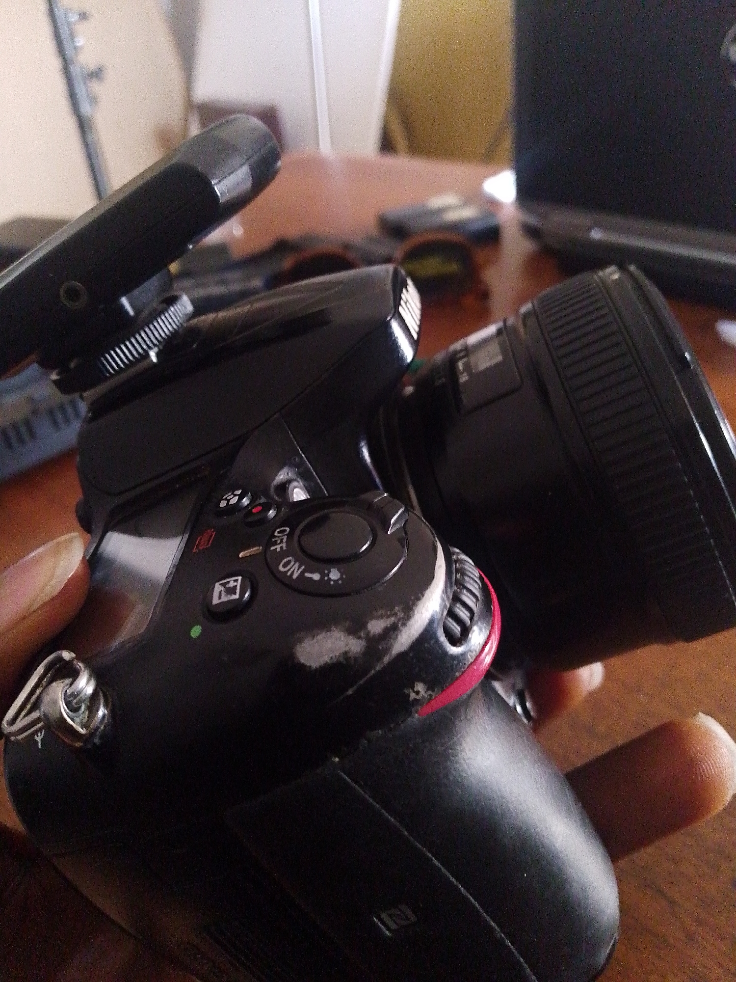 The image shows a close-up view of a black digital camera held by a person's hand. The camera has a lens attached and a flash unit mounted on top. Several buttons and dials are visible on the camera body, including an "ON" and "OFF" switch. Some wear and tear are visible on the camera's surface, particularly a scuff mark on the side. The background is blurred, suggesting the focus is on the camera. Hints of a wooden table and a laptop are visible in the background, implying the camera might be on a desk or table in an indoor setting. The lighting appears to be artificial or from an indoor source, as there are no strong shadows indicating natural daylight. No specific time of day, weather conditions, or explicit emotions are discernible from the image. There is no visible text in the image.