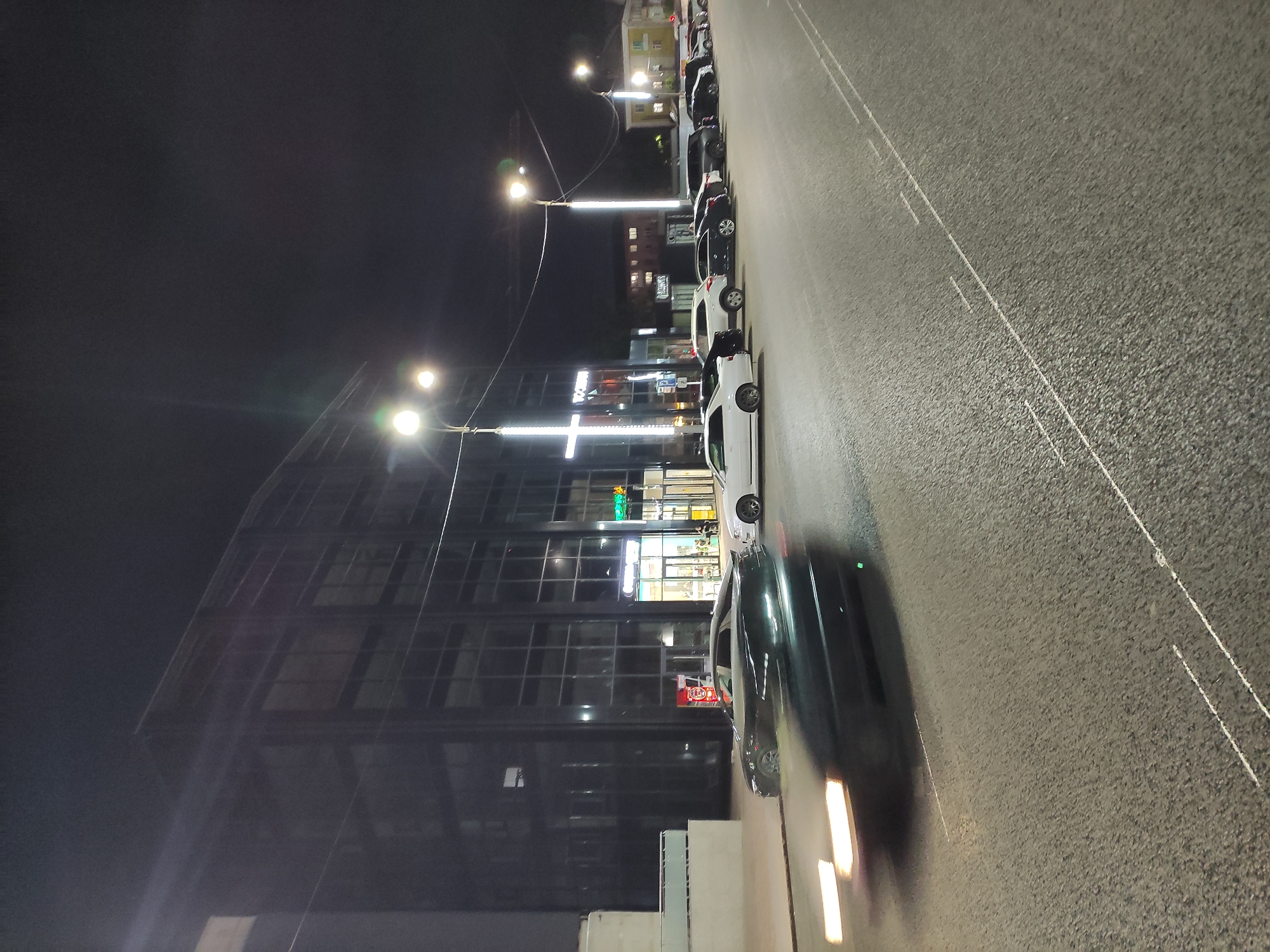 The image shows a street scene in Pavlodar, Kazakhstan at night. Several cars are parked along the side of the road. The street is illuminated by streetlights. On the left is a modern building with many windows. There are shops and businesses visible inside the building. The pavement appears to be asphalt. The night sky is dark.