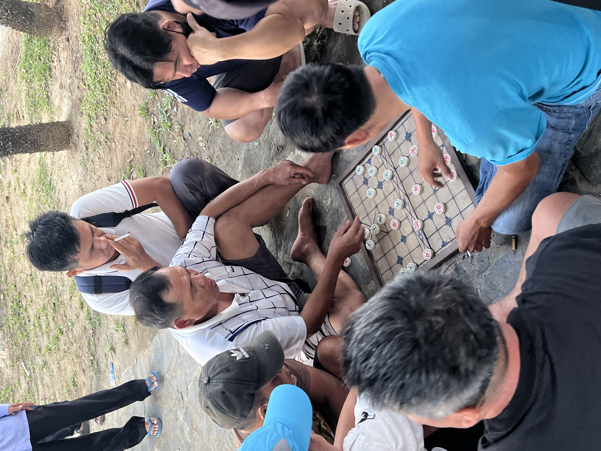 This image captures an informal gathering of approximately eight men engaged in a game of Xiangqi (Chinese chess) outdoors in Bắc Nha Trang, Vietnam. The scene unfolds on a patch of ground that appears to be a mix of dirt and rough concrete, with sparse grass and tree trunks visible in the background, suggesting a park or a casual public area.
At the center of the activity is a wooden Xiangqi board with circular playing pieces, each marked with distinct Chinese characters. Two men are actively playing, positioned on opposite sides of the board. One player, wearing a blue t-shirt and jeans, is seen leaning over the board, hand poised near the pieces, indicating he is either making a move or contemplating his next strategy.
The other men form a circle around the players, intently observing the game. Their attire is casual, including t-shirts, polo shirts (one with black and white stripes), shorts, and pants. One man, wearing a white polo shirt with a backpack, is smoking a cigarette. Several men are sitting or squatting on the ground; some are barefoot, while others wear flip-flops or sandals. A black Adidas baseball cap is visible on one man's head, and a small plastic water bottle lies on the ground near another observer.
The natural light indicates it is daytime, likely under an overcast sky or in a shaded spot, as there are no harsh shadows. The overall mood is one of focused concentration and relaxed social interaction, typical of a casual community gathering centered around a game.