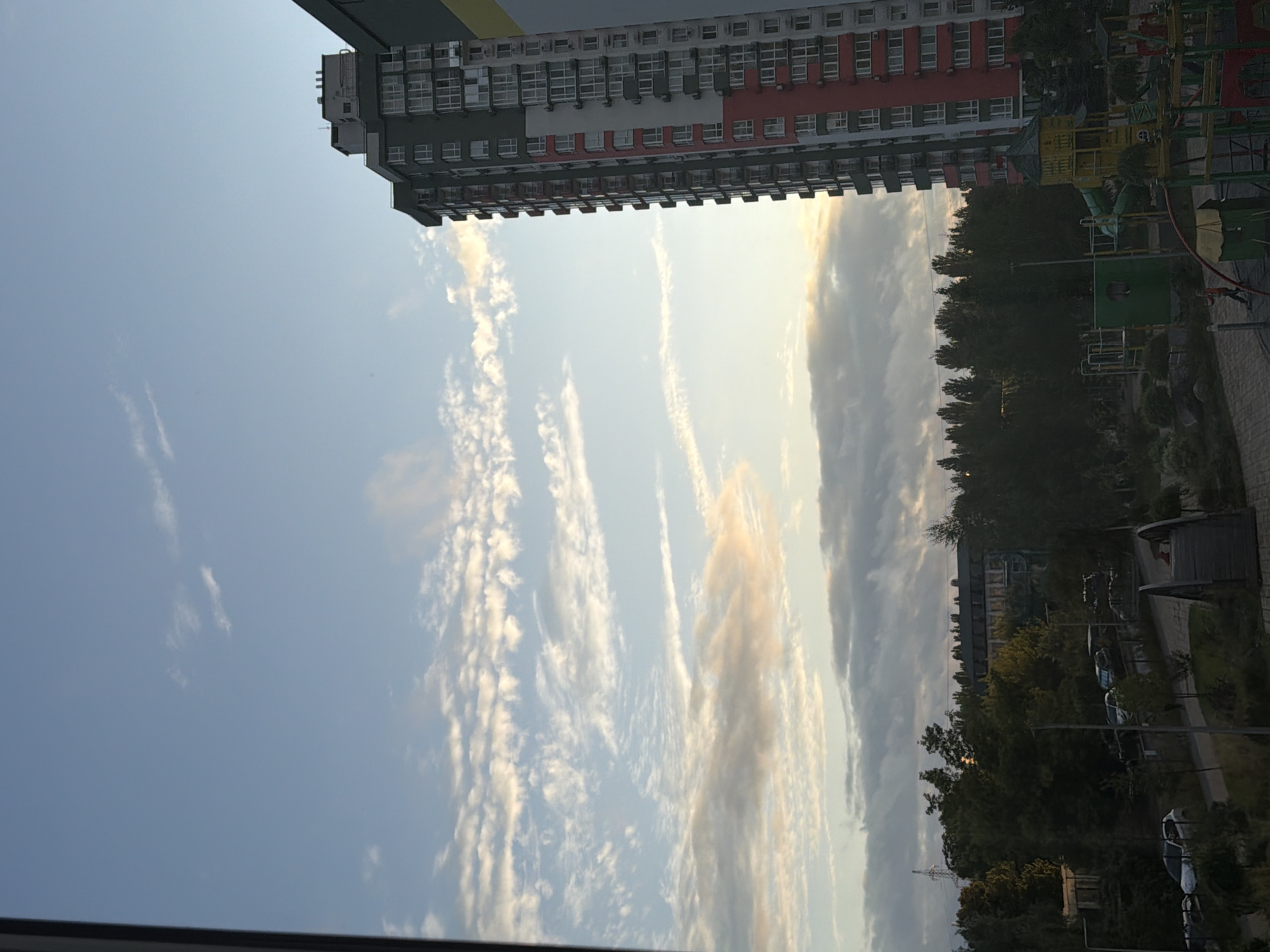 The image depicts a view from a window overlooking a residential area in Kyiv, Ukraine. A tall, modern apartment building with red and green accents dominates the right side of the frame, its facade punctuated by numerous windows. The sky above is a mix of blue and scattered clouds, with some clouds tinged with the warm hues of sunrise or sunset. Below the apartment building, a playground with colorful structures is visible, suggesting a family-friendly environment. Further into the distance, a dense line of trees separates the foreground from more buildings and infrastructure, including what appears to be a power line pylon. The lower part of the image shows a paved pathway, parked cars, and some landscaping, indicating a typical urban setting.