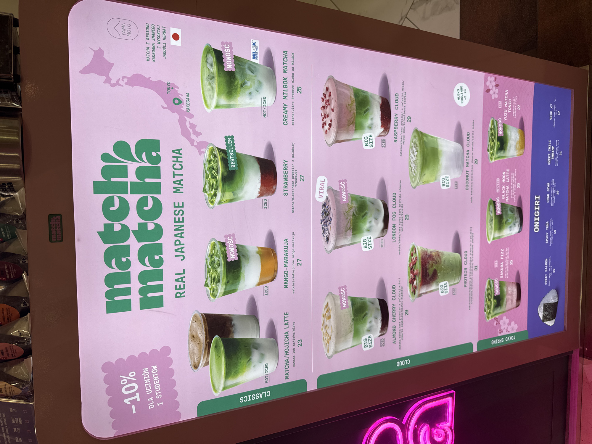 The image shows a brightly lit menu board for a matcha and onigiri shop. The brand name "matcha matcha" is prominently displayed in green lettering against a pink background. The board is organized into sections labeled "CLASSICS", "CLOUD", and "TOKYO SPRING". Each item on the menu is displayed with a corresponding image and price. The menu includes various matcha drinks, some with added flavors like hojicha, mango-marakuja, strawberry, and raspberry. There are also "cloud" drinks with flavors like almond cherry and coconut. Onigiri flavors, such as sweet salmon and spicy tuna, are also listed. Promotions such as discounts for students are also visible. A map of Japan highlights the Kakegawa region, known for its matcha production. The image indicates that the shop is located in Warsaw, Poland.