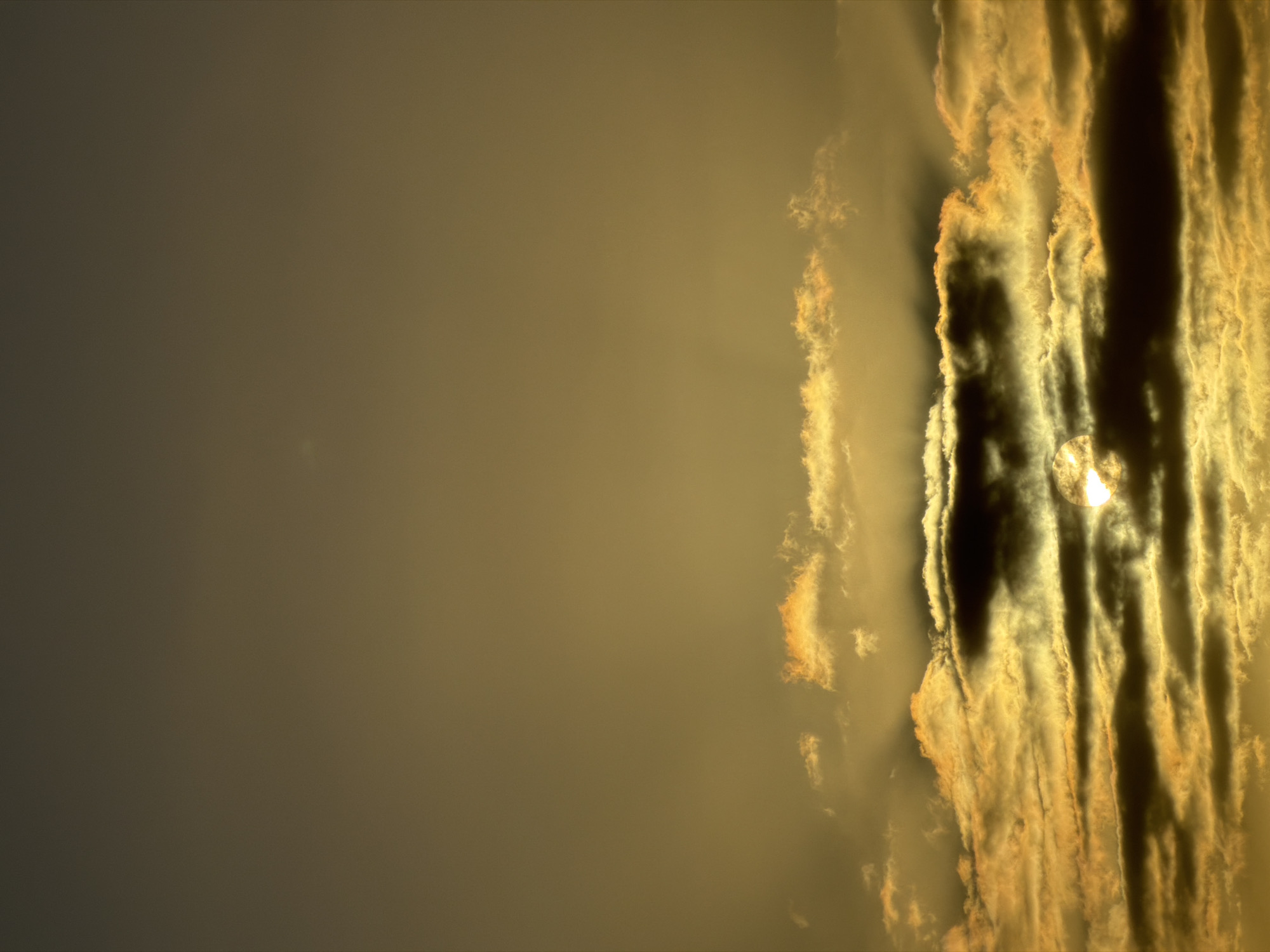 The image shows a sky with a strong yellowish-orange tint. The left side of the image is mostly a flat, uniform color, transitioning into a more dynamic area on the right side. Here, dense, striated cloud formations are present, with dark shadows contrasting sharply against the lighter, illuminated areas. The sun is partially obscured by the clouds, with a bright spot visible through the gaps. The overall atmosphere is hazy and atmospheric, suggesting either sunrise or sunset.