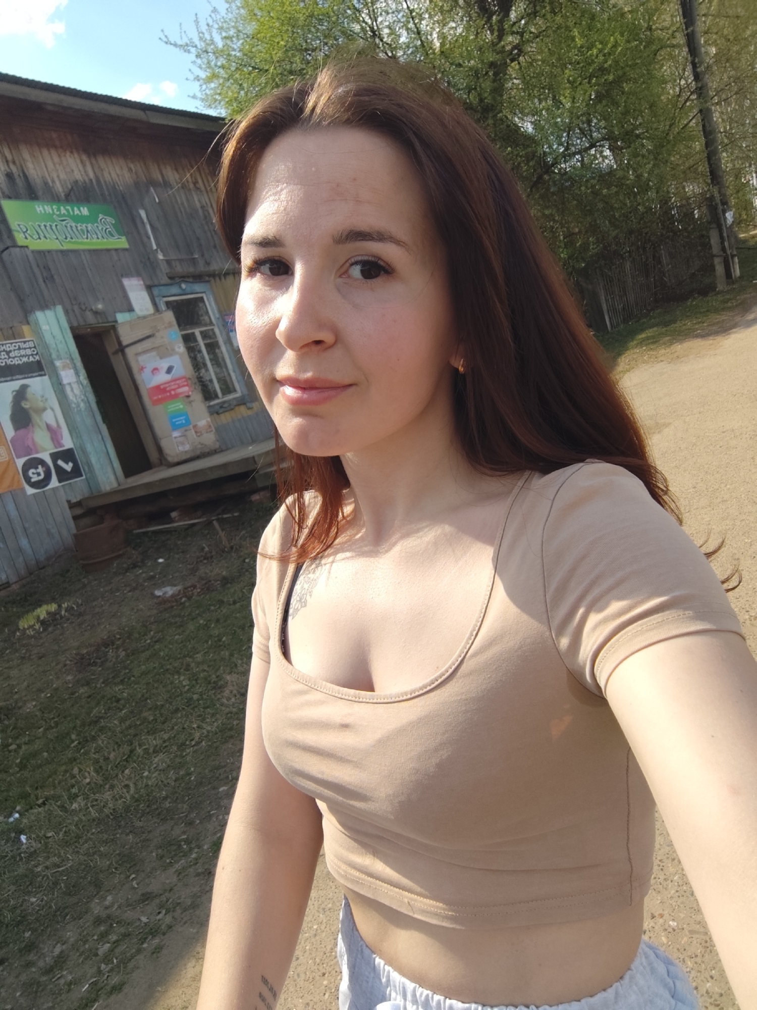 A woman with long brown hair is taking a selfie outdoors in the city of Фоки, Russia. She is wearing a tan crop top and gray sweatpants. Behind her is an old wooden building with a sign in Russian that reads "Магазин Канцтовары" (Stationery Store). There are posters and a cardboard box outside the entrance to the building. A dirt road runs alongside the building, and trees are visible in the background. The lighting suggests it is daytime.