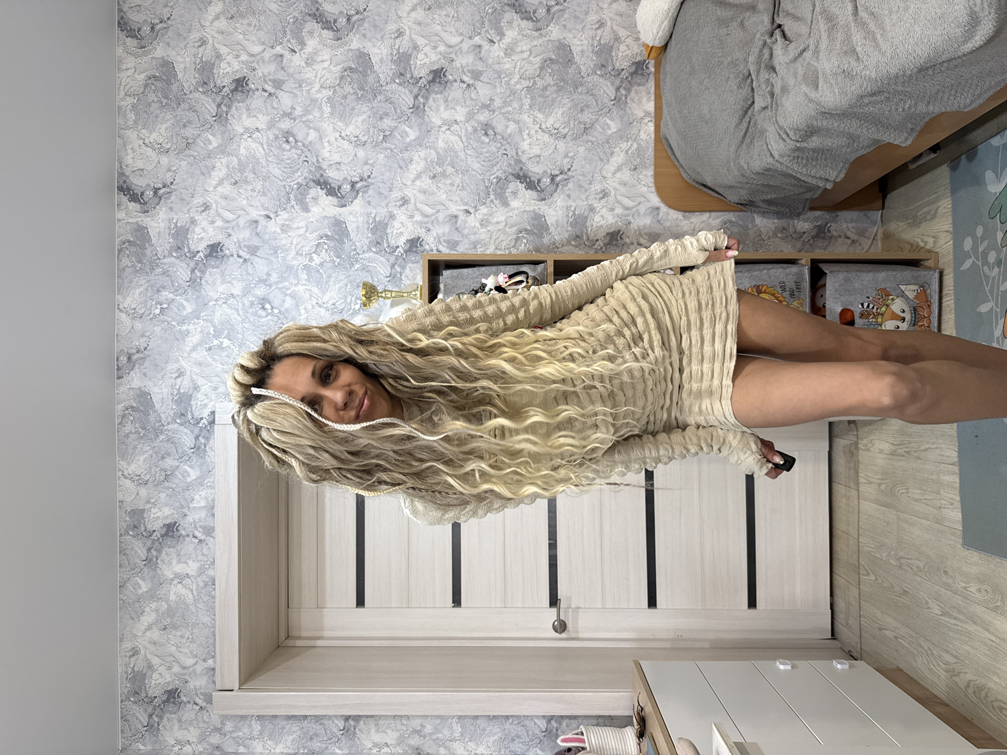 A woman with blonde, curly hair stands indoors in Шебунино, Russia. She wears a light-colored, textured dress with long sleeves. Behind her is a white door with black vertical inlays. To the left is a wall with a gray marble-like pattern. A bed covered with gray blankets is visible in the upper right corner. Below the bed, there is a shelf with gray storage boxes. The floor appears to be wood-patterned. In front of the door, a white cabinet with drawers is partially visible.