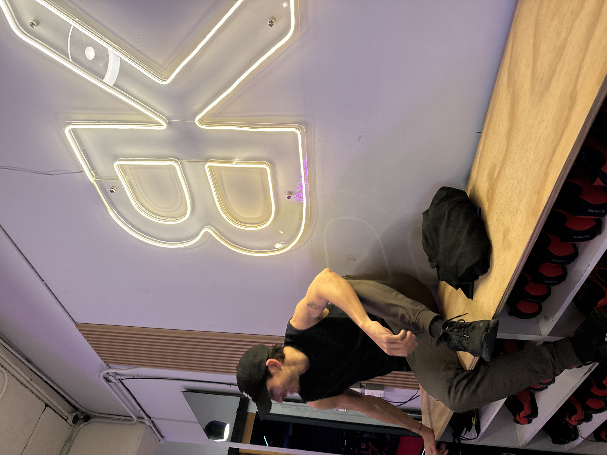 The image shows an indoor scene, apparently a gym or fitness studio, as suggested by the climbing shoes in the shelving system on the right. A man wearing a black t-shirt, dark pants, and a black baseball cap is standing and seems to be reaching for something. A black backpack is positioned near the shelving. The ceiling is white and features a large, stylized neon sign of two joined “B” letters. Based on the neon sign, the location is likely in Puebla, Mexico.