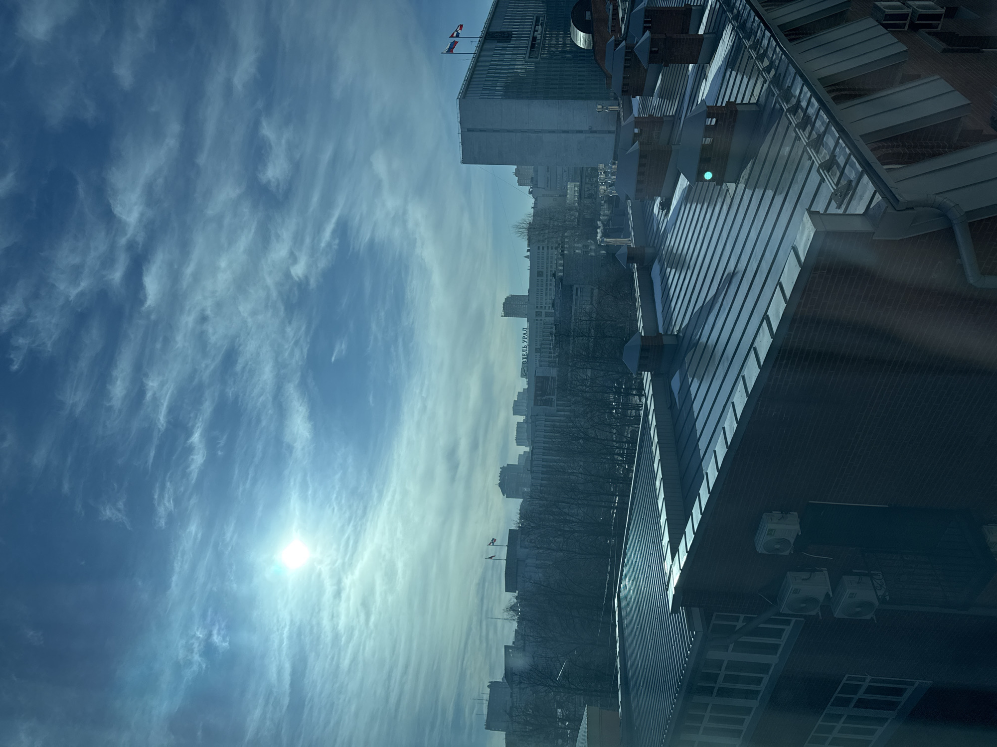 The image shows a daytime cityscape in Perm, Russia, viewed from an elevated position within a building. The sky is light blue with streaky cirrus clouds. The sun is visible as a bright spot in the lower left corner of the frame.
Below the sky, a row of buildings extends across the horizon. Prominent among these are the Hotel Ural building with visible Cyrillic lettering and a taller skyscraper with Russian flags flying on its roof. Trees, mostly bare, are interspersed among the buildings.
In the foreground, the edge of the building where the photo was taken is visible. Details include a metallic roof, ventilation ducts, air conditioning units attached to a brick wall, and window frames. A green glare is seen.