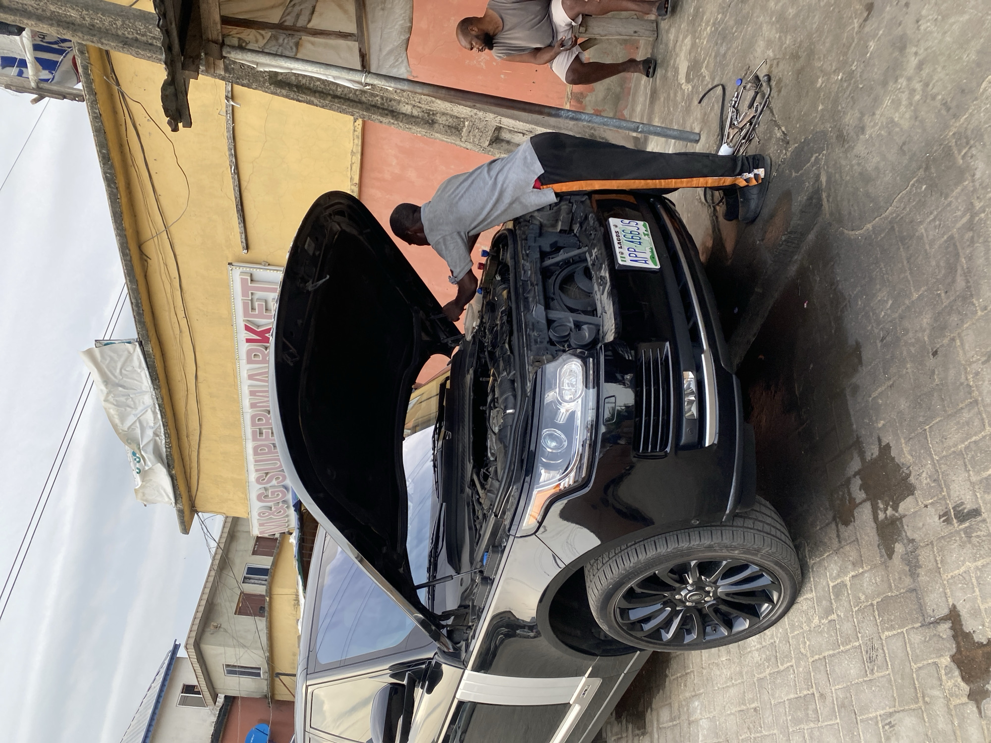 An outdoor scene in Ado, Nigeria, captures a car repair activity under an overcast sky. The ground is paved with concrete interlocking blocks, featuring some wet patches.
In the foreground, a black luxury SUV, likely a Range Rover, has its hood wide open, revealing the engine compartment. A man, presumably a mechanic, is actively working on the engine, bent over the front of the vehicle. He is wearing a light grey t-shirt and black pants with orange stripes. Various tools, including wrenches, are scattered on the ground near the car's front wheel. The car's license plate, "APP 466 JS," is visible on the front, with "LAGOS" inscribed below it.
In the mid-ground, another man with a beard is seated, observing the repair. He is dressed in a grey t-shirt and light-colored shorts. The background features several buildings; a prominent yellow-painted structure displays a partially visible sign that reads "M&G SUPERMARKET." Behind it, another building with a reddish-orange wall is discernible. Overhead, a makeshift structure of wooden beams, metal poles, and wires indicates an informal workshop setting. A satellite dish is visible on the roof of a distant building.
The scene depicts a focused daytime car repair in a local, open-air environment.