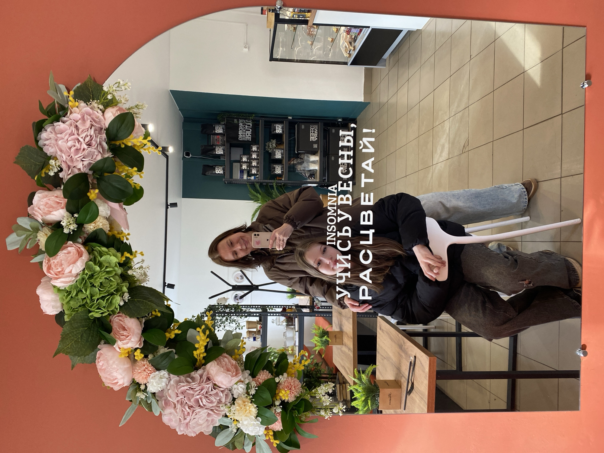 Here's a detailed description of the image:
**Scene:**
The image is a reflection in a decorative mirror hanging on an orange-painted wall, likely taken inside a retail or service-oriented establishment. The mirror is adorned with a floral arrangement of pink and green hydrangeas, roses, and yellow wildflowers along the left side.
**People:**
The reflection shows two females. One is holding a smartphone, presumably to take the picture. She is wearing a brown jacket. The other woman is in front and slightly below, wearing a black jacket and sitting on a white plastic chair. They appear to be posing or taking a selfie together.
**Objects:**
Besides the mirror and floral arrangement, the reflection shows retail shelving units stocked with products, possibly coffee or related items, suggesting the business is a cafe or shop. There is also a display case with items in it. A wooden table with utensils on it is also visible.
**Actions and Interactions:**
The primary action is the taking of a photograph using a smartphone. The two women are interacting, likely posing for the picture.
**Environment:**
The interior lighting appears to be standard commercial lighting. The flooring is tiled in a light color. The overall environment suggests a modern or trendy cafe/shop. Text on the mirror reads "Insomnia, learn to dream, bloom!"
The location is in the city of Chaikovsky, Russia.