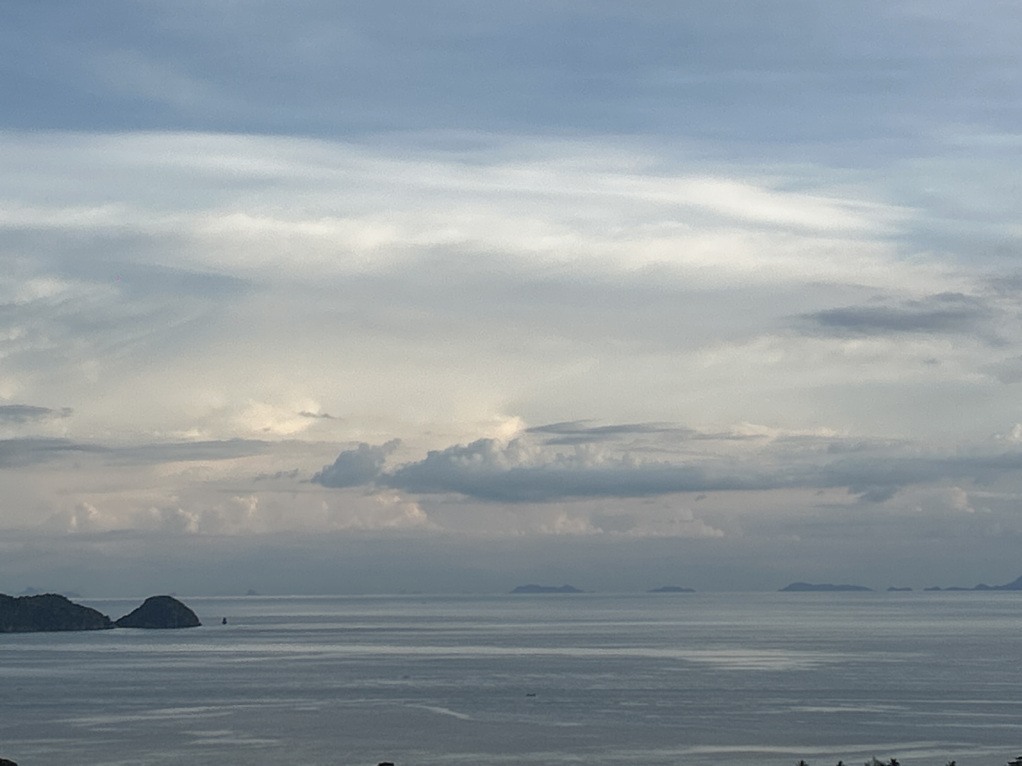 This media file depicts a serene seascape under a cloudy sky. The primary subjects are the vast ocean and the sky filled with layers of clouds, ranging from wispy streaks to larger, more defined formations. In the distance, several islands are visible as dark silhouettes against the horizon, hinting at a tropical or coastal location.
The scene appears calm, with gentle ripples on the water's surface. The overall mood is peaceful and tranquil, likely captured during either dawn or dusk due to the soft, diffused light. The sky's colors are muted blues, grays, and whites, suggesting overcast conditions or the transitional light of early morning or late evening.
A small, dark object, possibly a boat, is visible on the water in the middle distance, adding a touch of scale and activity to the expansive view. Two larger islands, one with a distinct conical shape, are closer to the left side of the frame, providing a more prominent land feature. The lighting and cloud formations suggest a slightly dramatic but beautiful sky, typical of coastal weather patterns. There is no visible text in the media file. The location context, ตำบลเกาะพะงัน, Thailand, places this scene in a well-known tourist destination known for its beautiful beaches and islands.