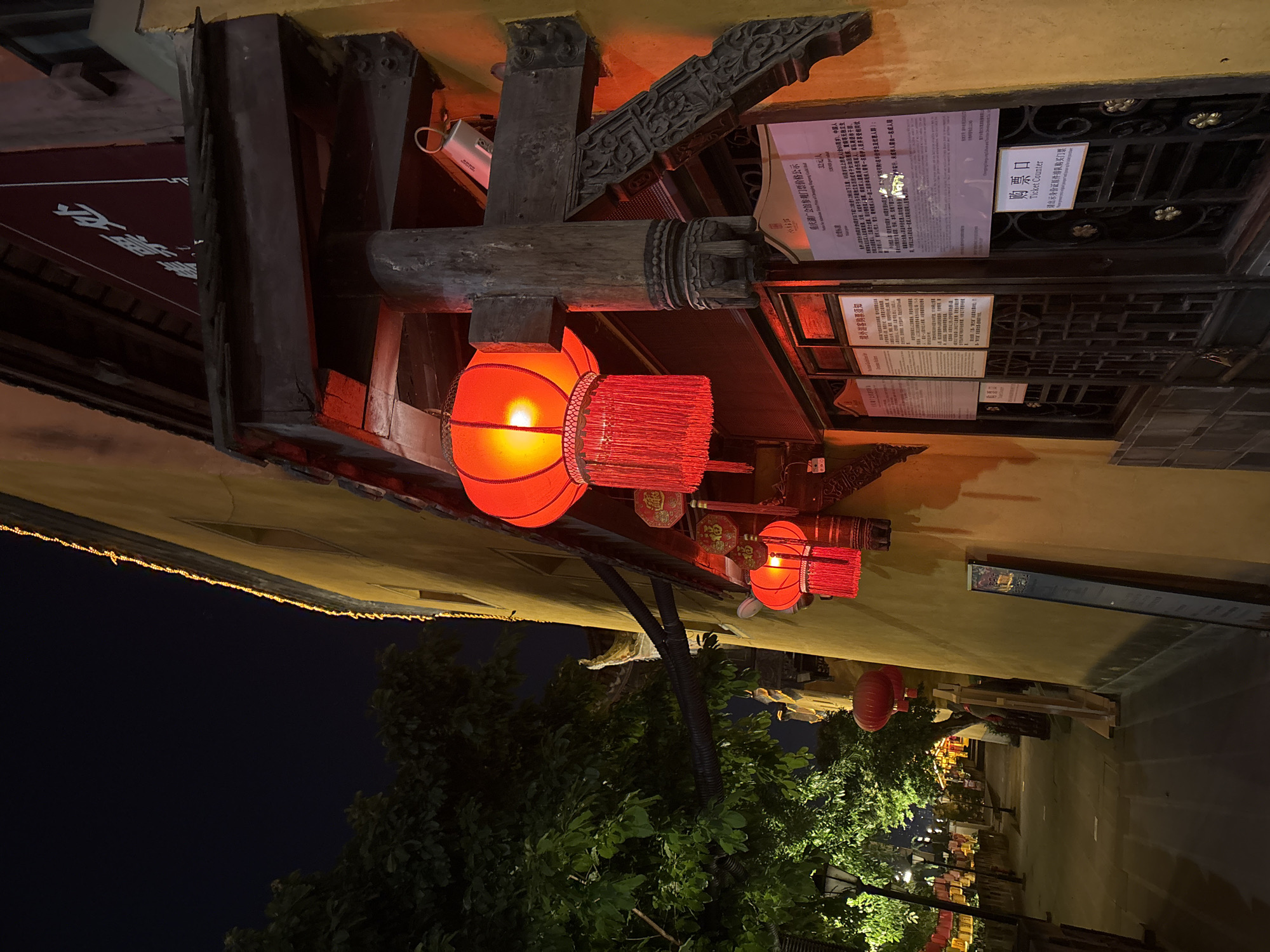 The image shows a view of an illuminated street in Yuzhong District, China, at night. Red lanterns are hanging from the eaves of buildings. A sign in Chinese and English, translated as "Ticket Counter," is visible on one of the buildings. The location is identified as Chongqing Huguang Guild Hall through other signs. Trees line the street, adding a touch of greenery to the urban setting. The sky is dark, indicating nighttime.