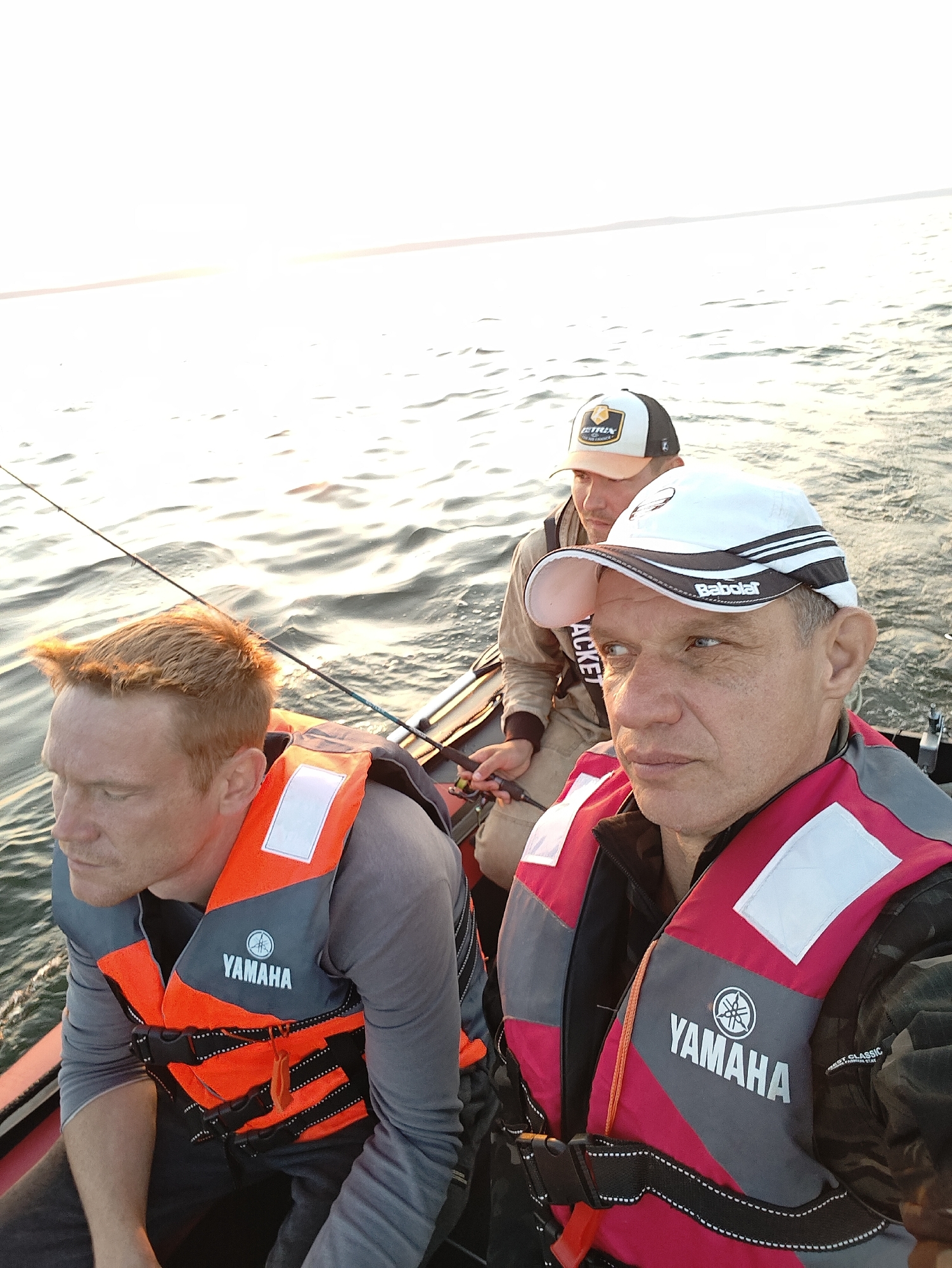 Three individuals are in a boat on the water, seemingly engaged in fishing. The person in the foreground, on the right, is wearing a white baseball cap with black and white stripes and the "Babolar" logo. They are also wearing a red and gray life vest with the "YAMAHA" logo visible. Their gaze is directed to the left of the frame. To the left of this individual, another person is visible, wearing an orange and gray life vest with the "YAMAHA" logo. This person has reddish-blonde hair and is looking down and to the left. In the background, a third individual is wearing a white and brown baseball cap. The boat appears to be in motion, with ripples on the water surface. The overall lighting suggests it is either early morning or late afternoon, with a bright, hazy sky. The scene is consistent with outdoor recreational activity, specifically fishing, on a body of water. Given the information, this scene could be on the Kama River or Votkinsk Reservoir near the city of Chaykovsky, Russia, a region known for its aquatic sports and fishing opportunities.