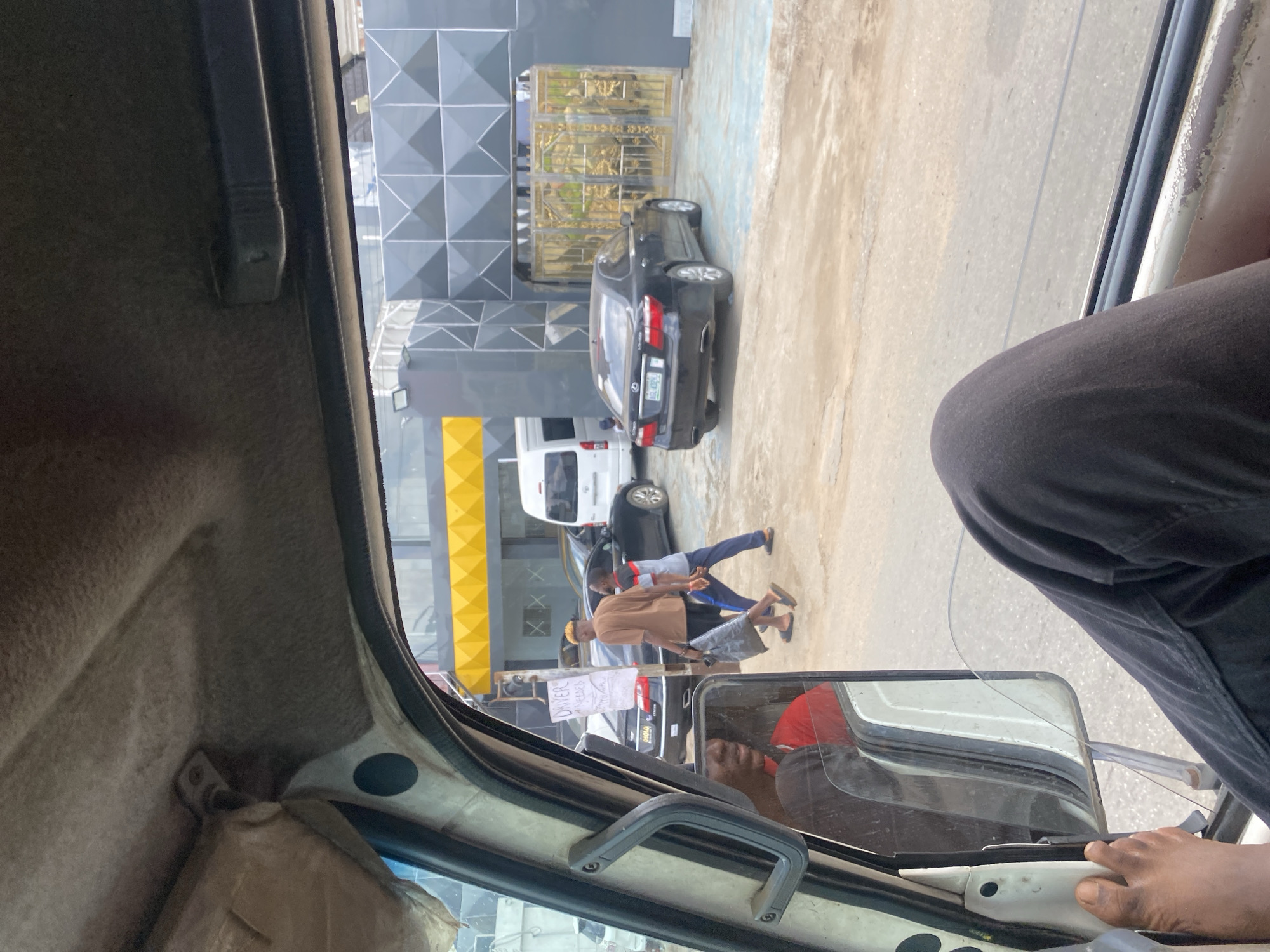 This media file captures a street scene in Idimu, Nigeria, from the perspective of someone inside a vehicle, likely a commercial bus or similar. The image is taken during the daytime under bright, clear weather conditions.
In the immediate foreground, parts of the vehicle's interior are visible, including the light-colored ceiling liner, a dark grab handle, and a side mirror. The reflection in the side mirror shows a person in a red shirt, likely the driver. The leg and right foot of a person, dressed in black trousers, are also visible on the right side of the frame, resting near the vehicle's door or dashboard.
Outside the vehicle, a bustling street scene unfolds. To the left, a modern building with a distinctive gray and black geometric facade stands, partially obscured by a golden ornate gate. Below this, another building with a bright yellow patterned section is visible. A black sedan, bearing the license plate "AGL 698GG," is parked on the dusty ground outside the buildings. A white van is also parked further down the street.
Several people are walking on the pavement. One individual in a brown shirt and light-colored pants is walking towards the left, carrying a dark bag. Another person in a blue shirt and dark pants walks alongside them. Near these pedestrians, a handwritten sign is displayed on a stand. The visible text on the sign clearly reads "DRIVER NEEDED," followed by partially obscured words and a phone number, suggesting a job advertisement.
The overall scene depicts a typical urban or suburban street environment in Nigeria, characterized by vehicle traffic, pedestrians, and commercial activity alongside distinctive architecture.