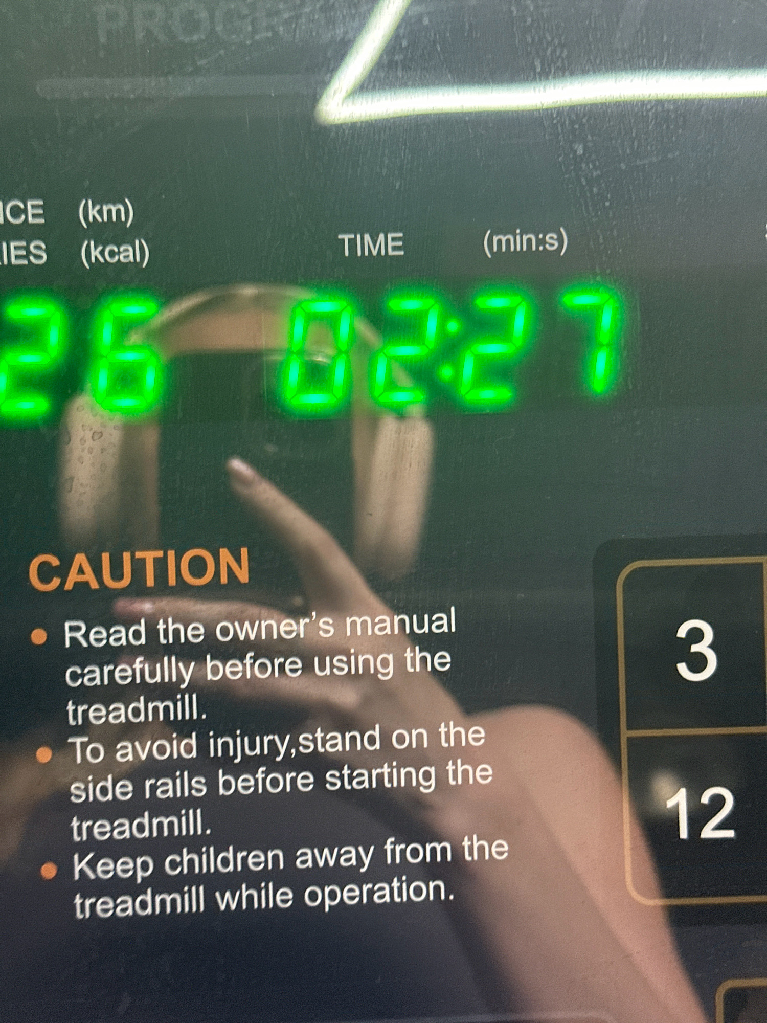The image shows a close-up of a treadmill display and a portion of its console. The display shows green LED digits indicating "26" for distance in kilometers, "02:27" for time in minutes and seconds. To the right, there are two white number displays, one showing "3" and the other showing "12". Below the distance and time, there are caution instructions printed in white text on a dark background. These instructions include points about reading the owner's manual, standing on side rails, and keeping children away. The background is dark and slightly blurred, with a faint outline of what appears to be a person's arm and hand. The overall impression is of a fitness device in operation, likely in an indoor setting. There is no visual information in the image that could place it in Lalitpur, Nepal, or any specific city.