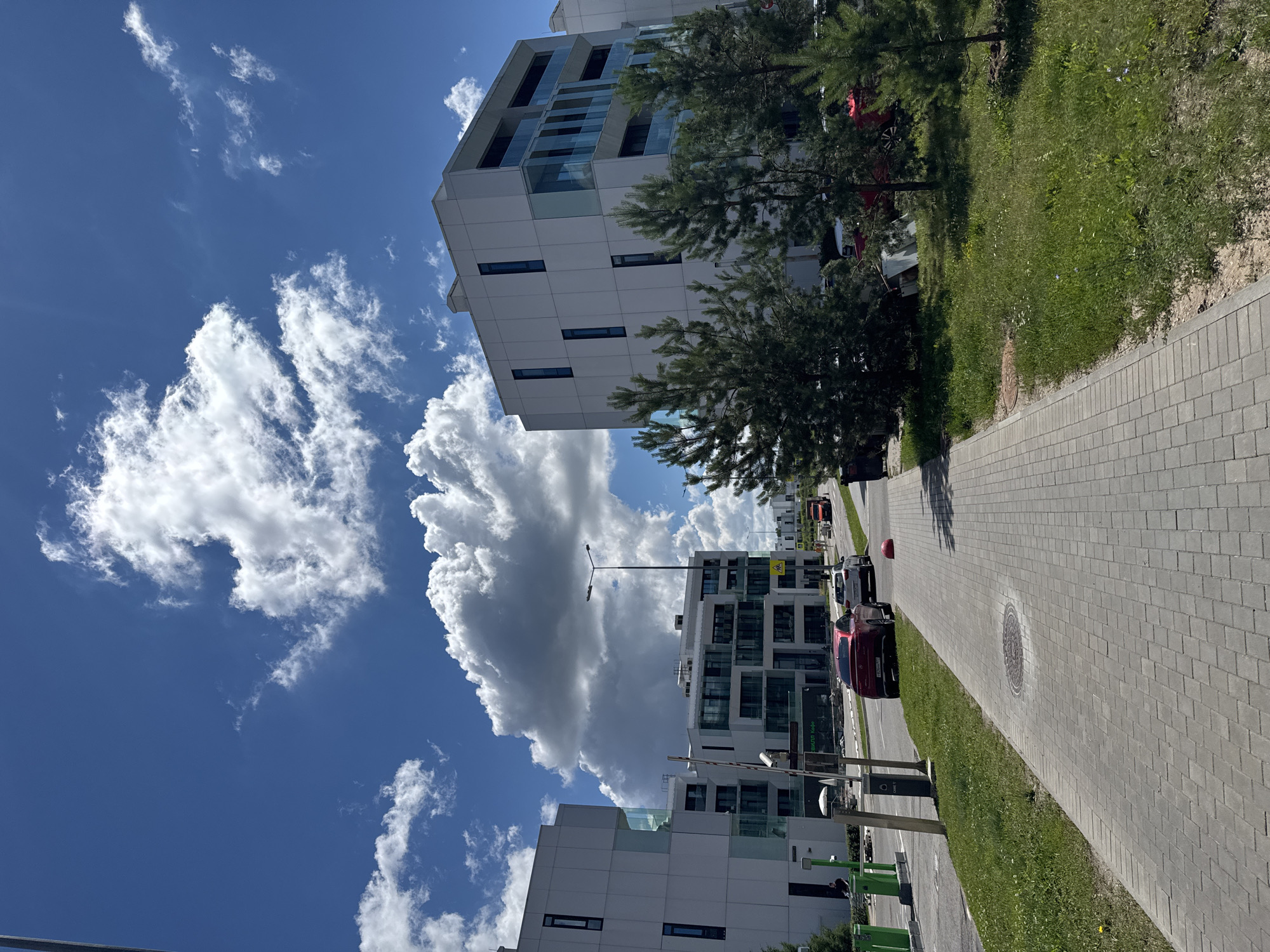 The image depicts a modern residential complex on a clear, sunny day with a vibrant blue sky dotted with fluffy white clouds. The architecture features clean lines, white facades, and extensive use of glass balconies, suggesting a contemporary urban development. Two multi-story buildings dominate the scene, with one on the left and another, larger one on the right.
In front of the buildings, a paved pedestrian walkway runs parallel to a street. Several cars are parked or driving on the street, including a red sedan and a white SUV. Green grass and mature pine trees line the walkway and the street, adding a touch of nature to the urban environment. A lamppost stands near the street, and a security camera is visible near the entrance of the building on the left. The overall impression is one of a well-maintained and modern living space, likely in a suburban or newly developed area of a city. While the image does not explicitly state its location, the presence of Cyrillic text on a sign visible in the image ("Свежий Кофе") indicates a location in a Russian-speaking country. Given the architectural style and general urban setting, it is plausible to be in a metropolitan area like Moscow, Russia.
