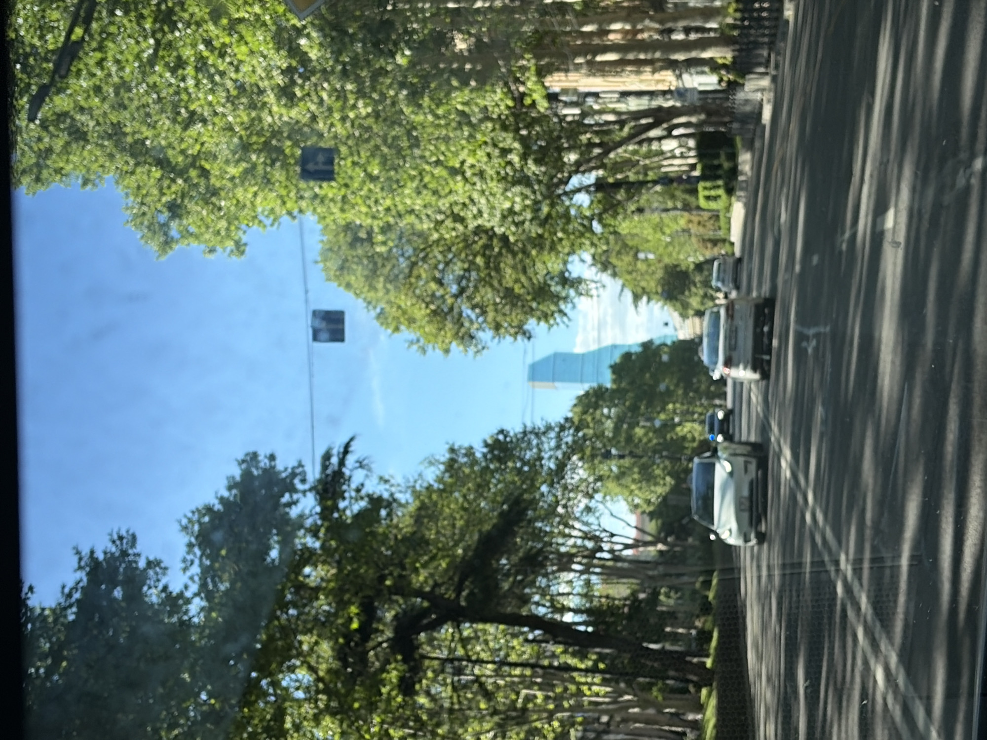 The image captures a street in Tbilisi, Georgia, lined with trees and buildings. In the background, a modern skyscraper can be seen. Cars are driving on the road. Two road traffic signs are also visible, suspended from the power lines.