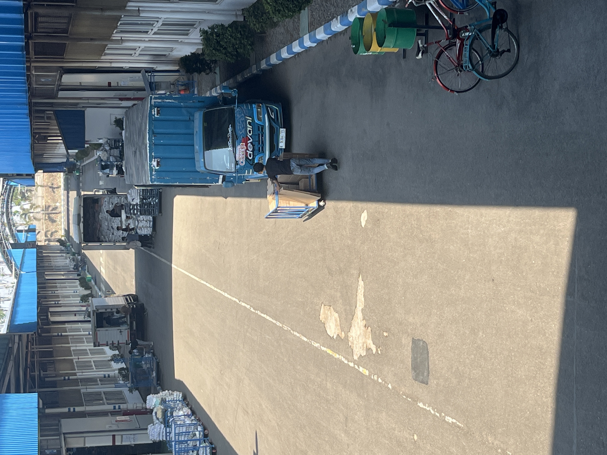 This image depicts an outdoor scene at a logistics or industrial facility in Pelang, Indonesia. The foreground is a paved asphalt area with a white line marking a lane. On the right side of the paved area, there are three large cylindrical trash bins, two yellow and one green. Next to the bins, two bicycles are parked: one red and white, and one light blue.
In the middle ground, a young man wearing a black t-shirt and blue jeans is pushing a wooden cart with several large cardboard boxes. He is walking towards a bright blue truck with the word "devani" in red lettering on its side. The truck has its back door open, suggesting loading or unloading activities. The truck has a license plate that reads "K 8784 FC".
Further in the background, on the left side of the image, are multiple rows of blue metal carts laden with white bags or packages. These carts are arranged neatly along the side of a building. The building has a blue roof and a series of large windows or doors. On the right side of the background, another truck, this one white, is parked with its back open and several people are visible near it, also seemingly engaged in loading or unloading. The overall environment appears to be a sunny day, with shadows cast across the paved area. The buildings suggest a functional industrial or warehouse complex.