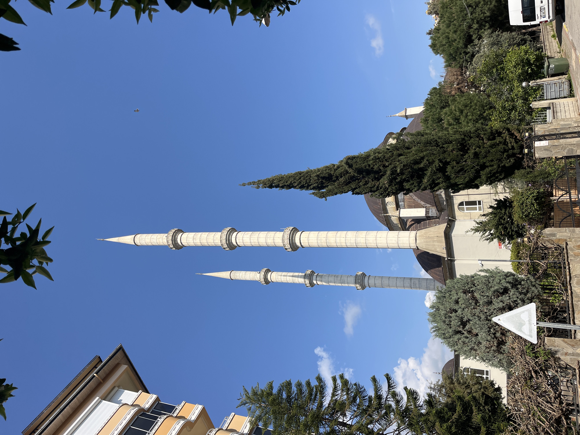 The image captures a low-angle view of a mosque in Alanya, Turkey, showcasing two minarets against a clear blue sky. The minarets are tall and slender, with circular sections and pointed tops. The mosque itself features a dome and is partially obscured by trees. The architecture is typical of Ottoman-style mosques. Various trees and foliage surround the mosque. In the foreground, parts of nearby buildings are visible, including a modern building with a yellow facade. A bird is visible in the clear blue sky. The overall scene depicts a sunny day in a residential area of Alanya, showcasing the blend of modern and traditional elements.