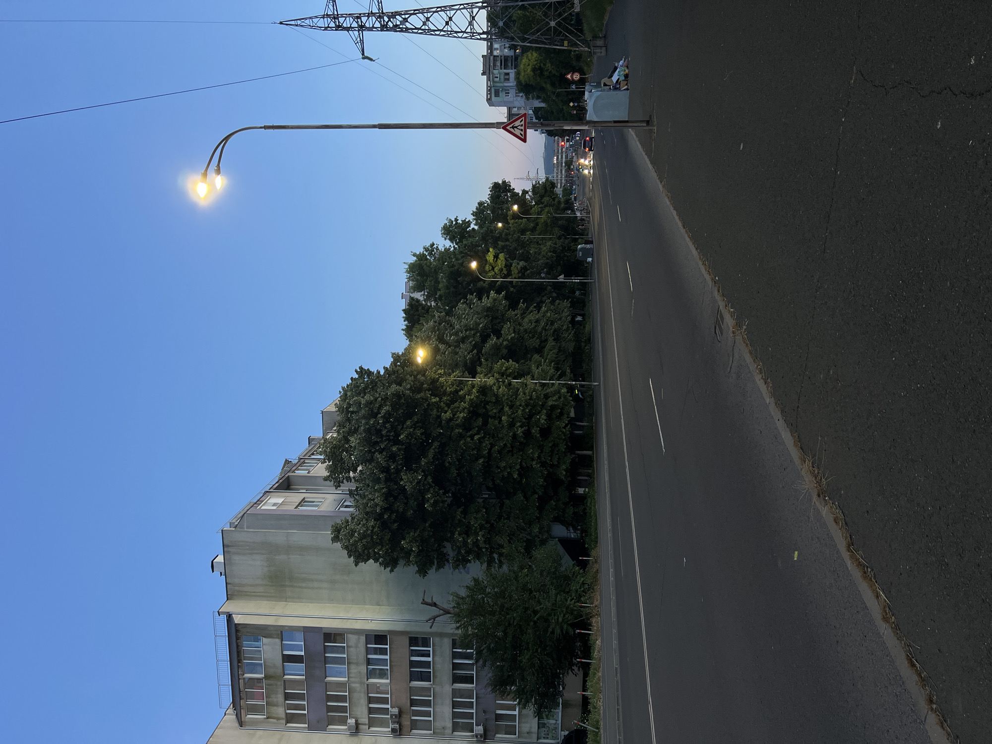 The image depicts a street scene in Burgas, Bulgaria, during twilight. The perspective is from the sidewalk, looking down a wide, multi-lane road. On the left side of the road is a tall, Soviet-era apartment building with multiple windows. Adjacent to the building are mature trees that line the street. On the right side of the road, there's a large metal pylon supporting high-voltage power lines. Further down the road, several streetlights are illuminated, casting a warm glow. Two lanes of traffic are visible, with a few cars driving in the distance. A pedestrian crossing sign is prominently displayed on the right side of the road, near a trash can and some discarded items. The sky is a clear, deep blue, characteristic of dusk.