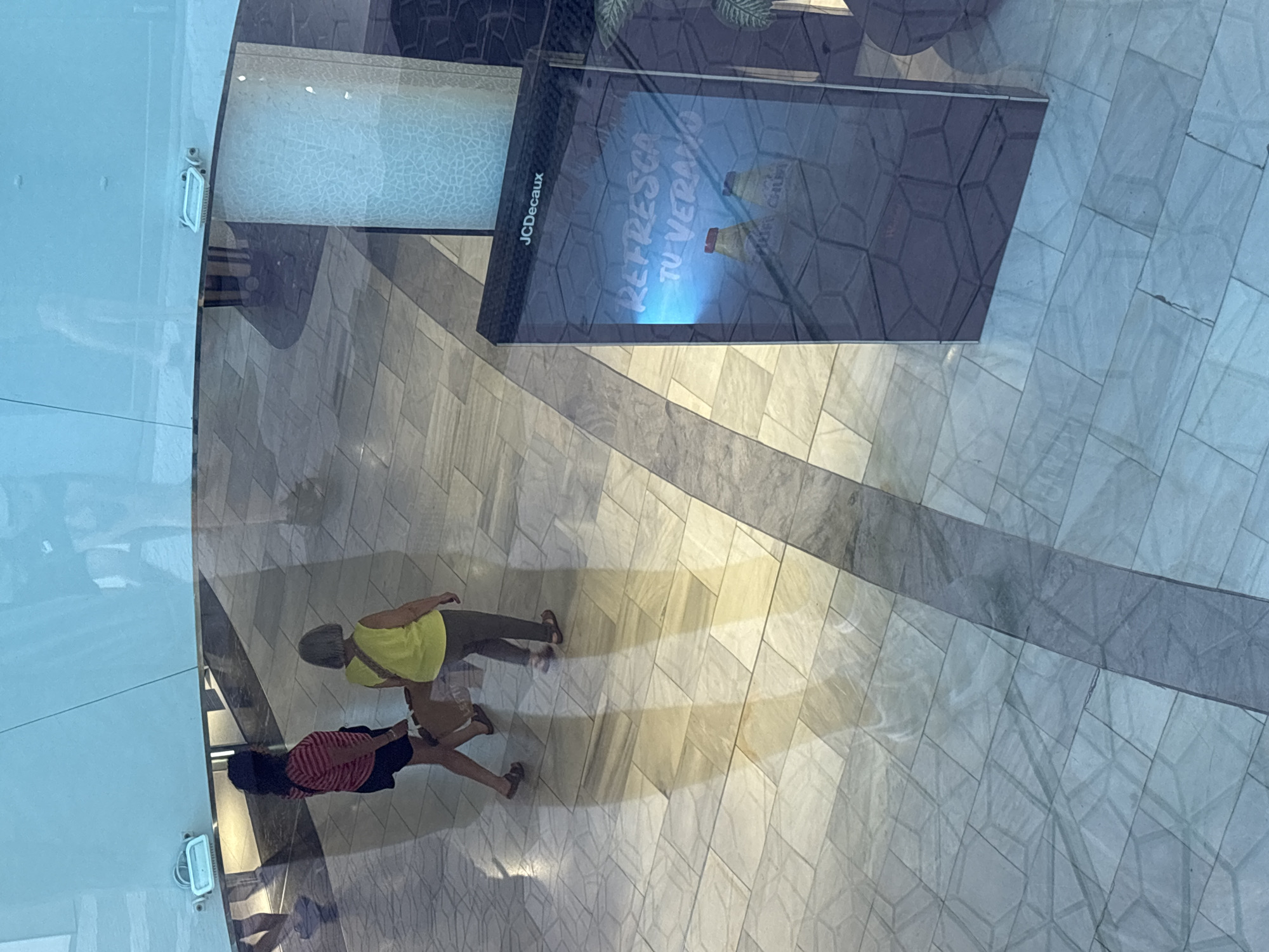 Captured from an elevated, slightly angled perspective looking down, the image depicts an indoor public space, likely a modern shopping mall or large building in Barcelona, Spain.
Two individuals are walking across a light-colored, patterned tiled floor. The person in the foreground wears a red and white striped t-shirt, dark shorts, and sandals. Ahead of them, a second person is dressed in a bright yellow top, dark pants, and sandals, carrying a light-colored shoulder bag. Both individuals cast faint reflections on the polished floor, which features a geometric design of light and dark grey rectangular tiles arranged diagonally. Overlaying parts of the floor, particularly on the right, is a subtle hexagonal grid pattern, possibly a reflection from above.
In the upper portion of the frame, a tall, rectangular digital advertising display is visible. The brand name "JCDecaux" is printed on its side. The screen shows an advertisement with the prominent Spanish text "REFRESCA TU VERANO" (Refresh Your Summer) in white, set against a blue background, accompanied by what appear to be two bottles. A green, leafy plant is partially visible near the top of the display.
The left side of the image is dominated by a semi-transparent, light blue surface, suggesting a glass balustrade or viewing platform, with a white light fixture attached. The overall lighting is bright and even, indicative of an indoor daytime setting. The people appear to be casually moving through the space, suggesting a routine daily activity.