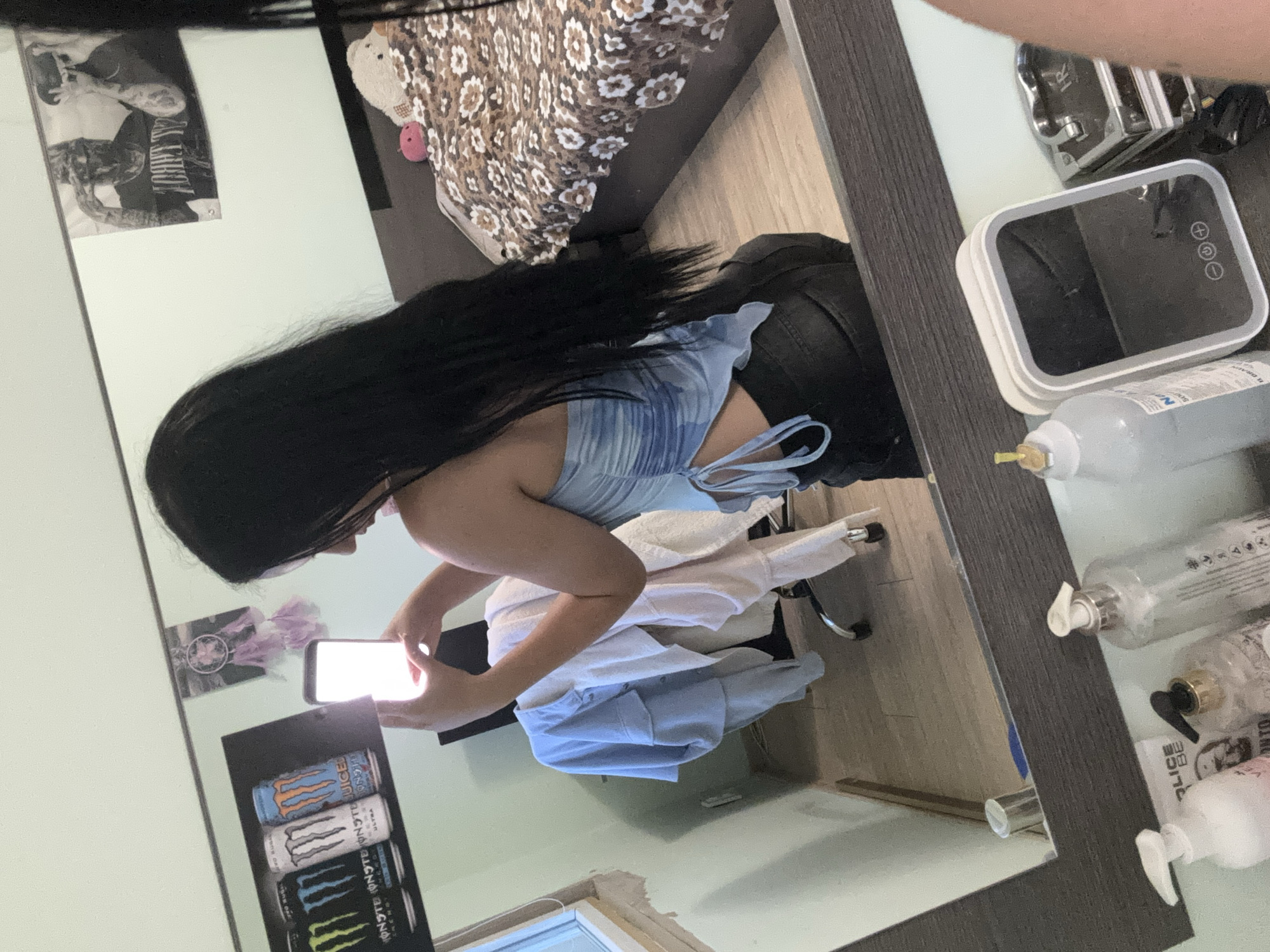 In a bedroom with light blue walls, a young woman with long black hair stands facing a mirror, taking a selfie with her phone. She is wearing a blue and white tie-dye crop top and black pants. The mirror reflects various items, including a bed with a floral comforter, a desk chair draped with clothes, posters on the wall, and a vanity with toiletries. The flooring is light wood laminate.