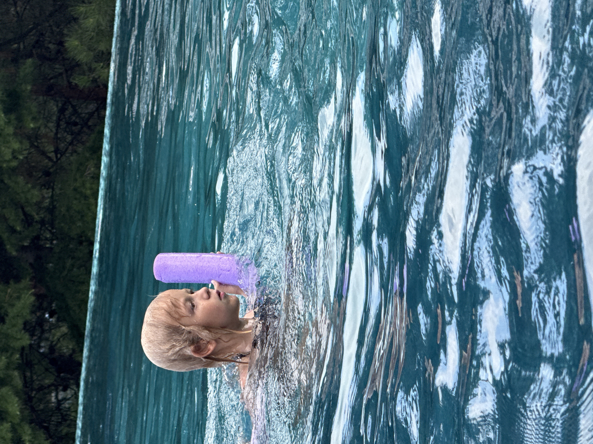 A young girl with blonde hair is swimming in a pool and holding a purple pool noodle. She is partially submerged in the water, which is a vibrant turquoise color and has ripples and reflections on its surface. The background shows a lush green forest and the edge of the pool. The scene appears to be set in an outdoor location, possibly a resort or private residence.